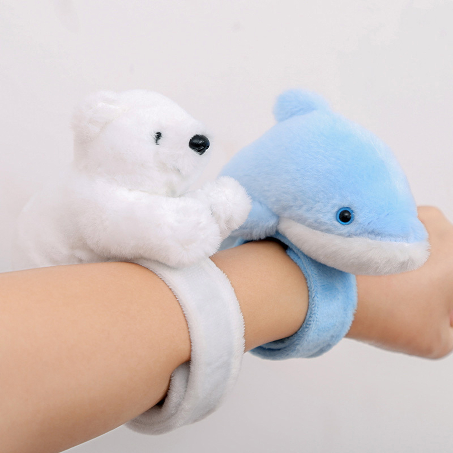 Una mano che indossa due braccialetti di peluche: uno con un orso polare, l'altro con un delfino, entrambi azzurri e bianchi, su sfondo bianco.