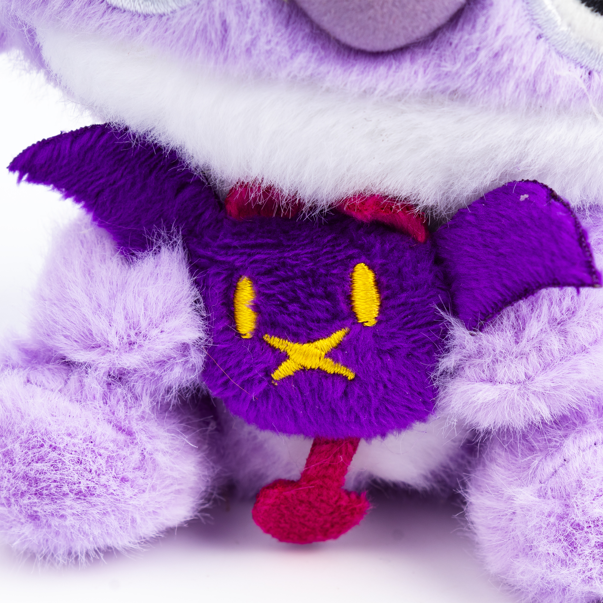 Un peluche viola con viso viola, croce e occhi gialli e dettagli rossi. Lo sfondo è bianco.