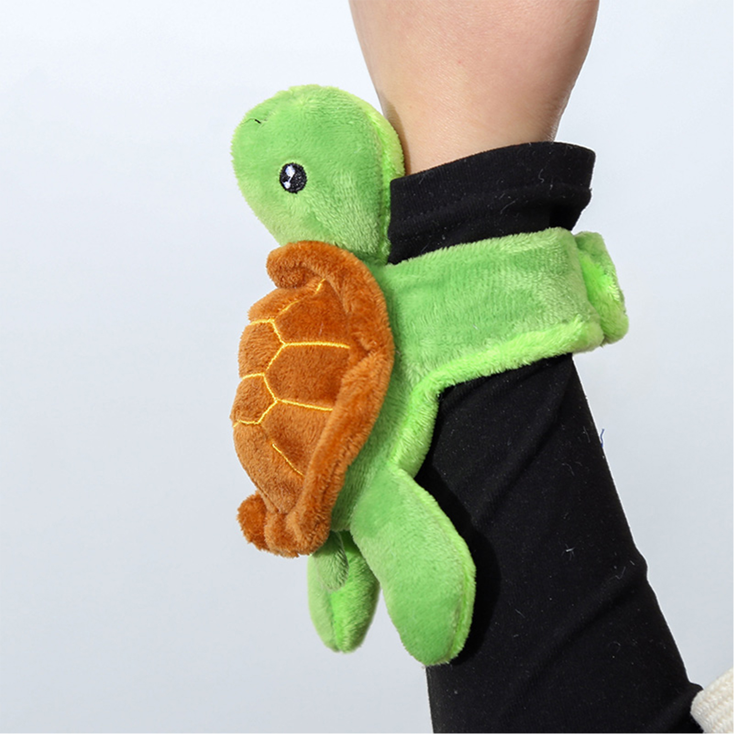 Un peluche a forma di tartaruga verde è attaccato a un polso. La tartaruga ha un guscio arancione ed è tenuta da una manica nera. Lo sfondo è bianco.