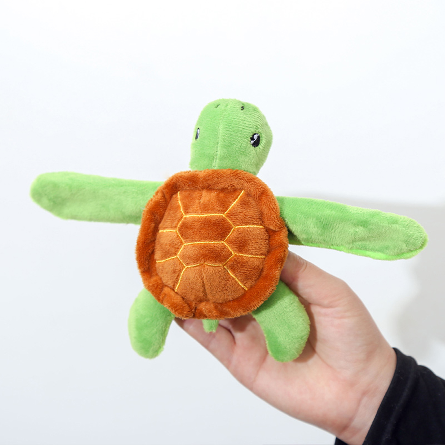 Un peluche a forma di tartaruga verde con guscio arancione è tenuto in mano su sfondo bianco.