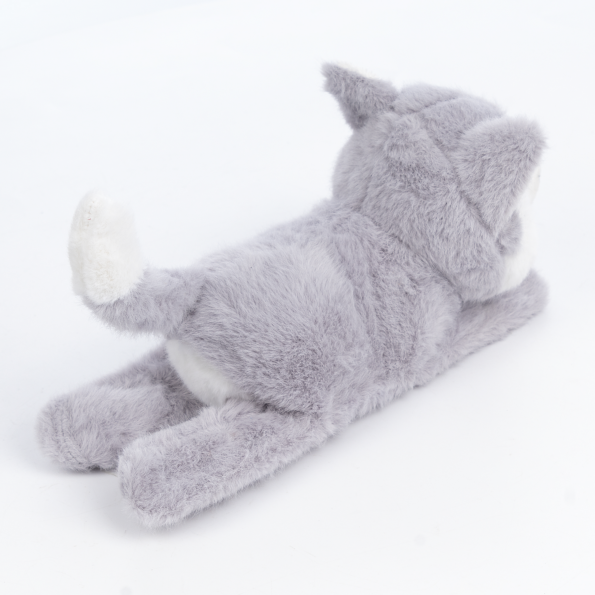 Un giocattolo di peluche grigio a forma di gatto è sdraiato su una superficie bianca. Il gatto è sulla pancia, si vedono la coda e le orecchie.