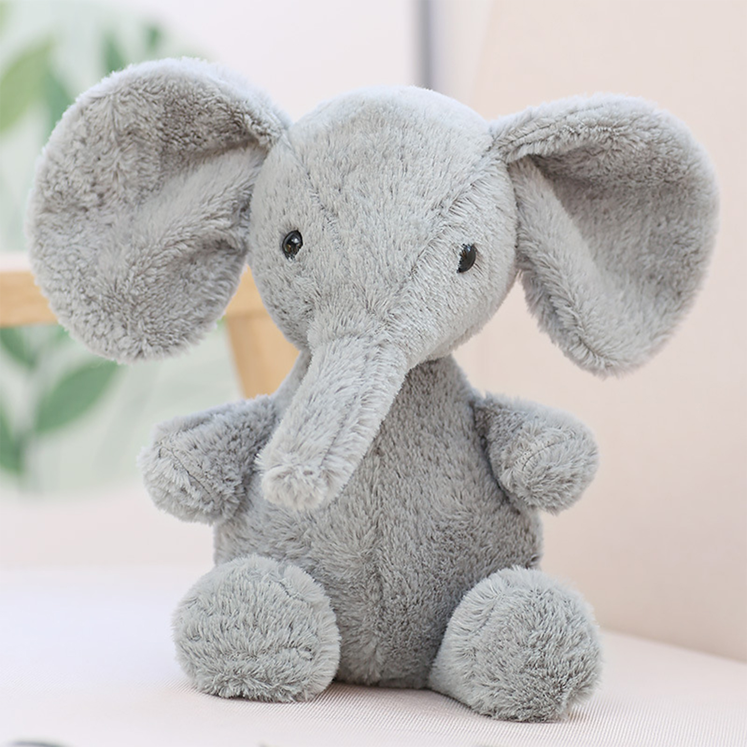 Un elefante di peluche grigio con grandi orecchie, una proboscide e piccoli occhi neri. È seduto su una superficie rosa con sfondo sfocato.