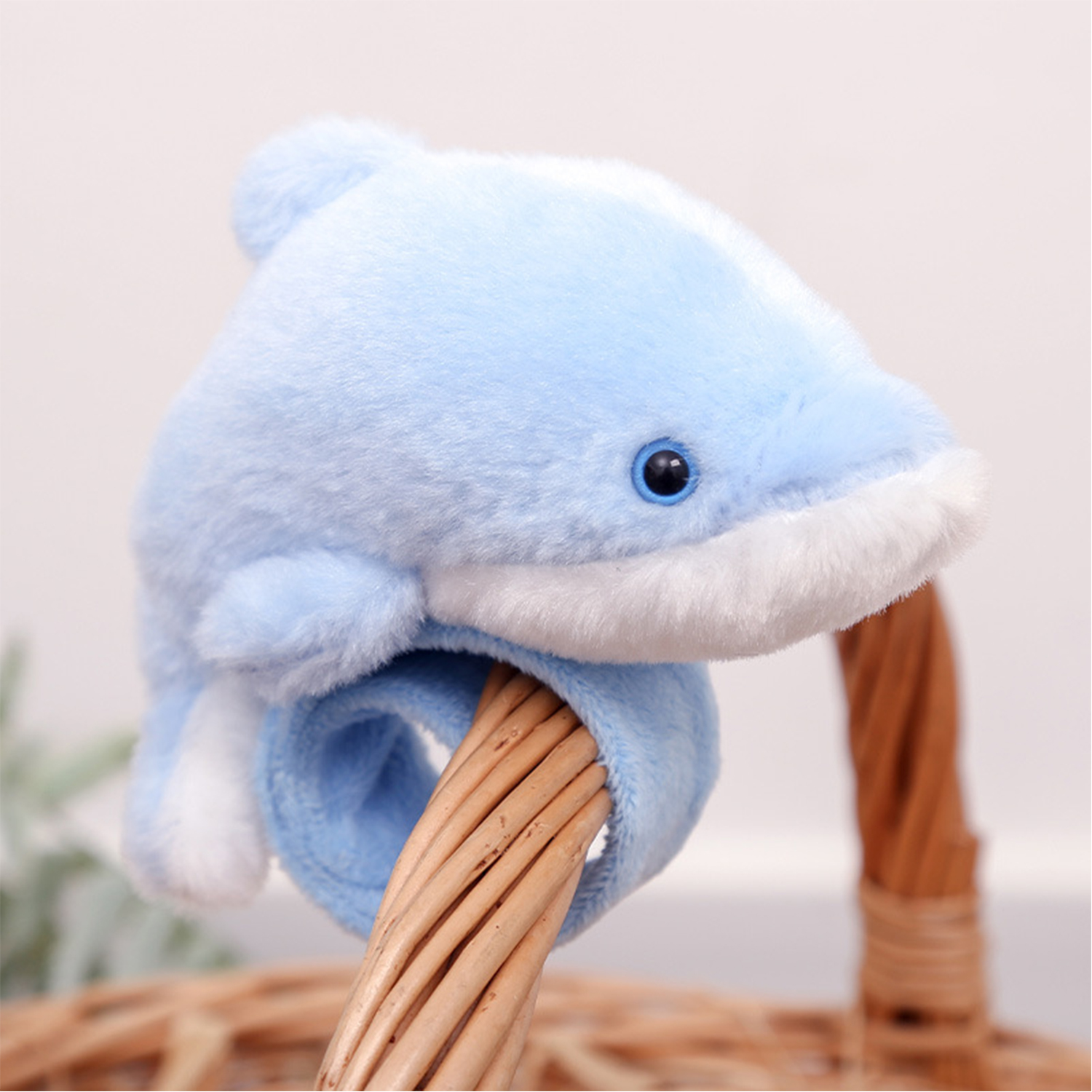 Un peluche a forma di delfino blu è appollaiato su un cestino intrecciato. Il delfino ha dettagli bianchi e un occhio scuro. Lo sfondo è sfocato.