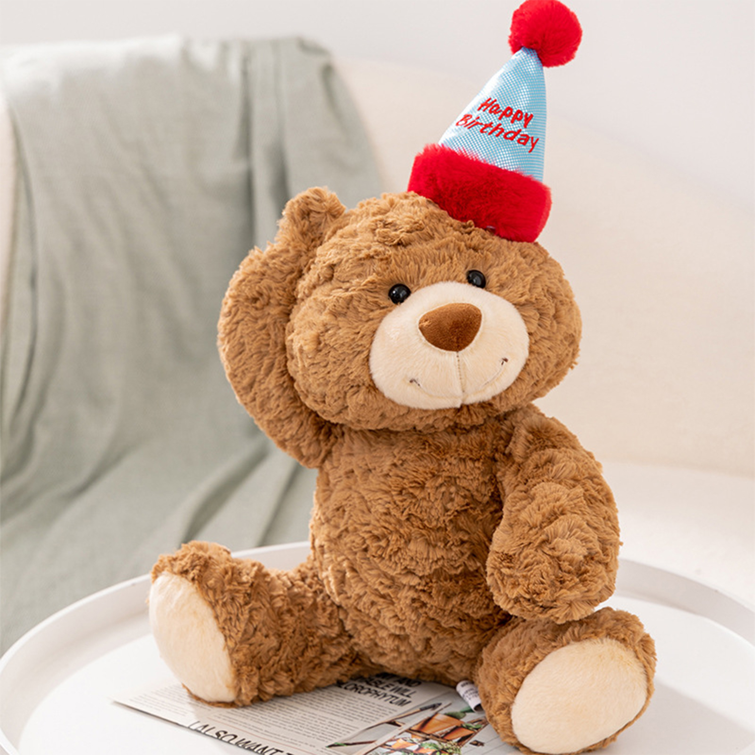Un orsacchiotto marrone con un cappello di compleanno siede su un vassoio bianco con un giornale. Una coperta verde chiaro sullo sfondo.