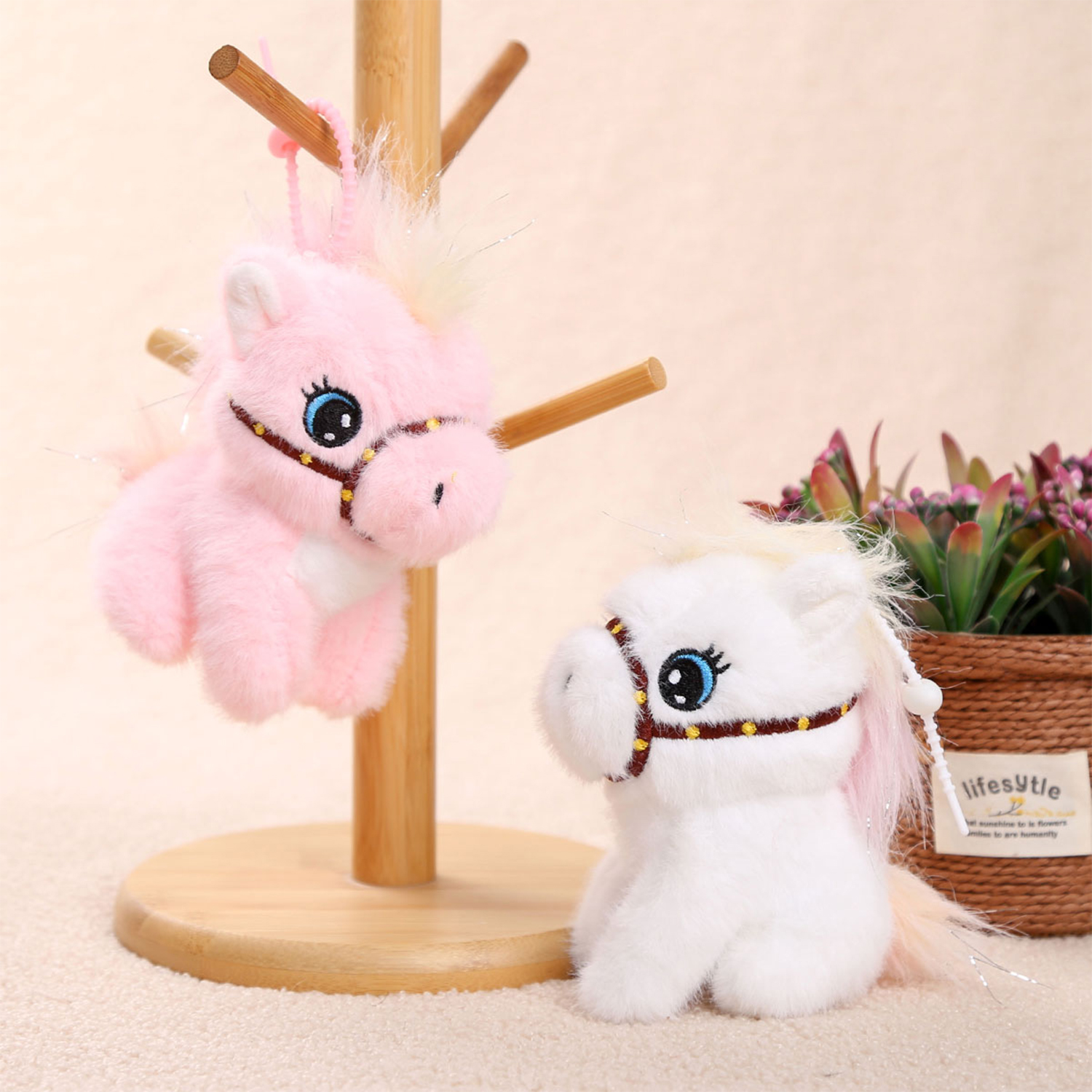 Due cavalli di peluche, uno rosa appeso, uno bianco seduto. Entrambi hanno finimenti marroni e volti con occhi blu. Supporto in legno sullo sfondo.