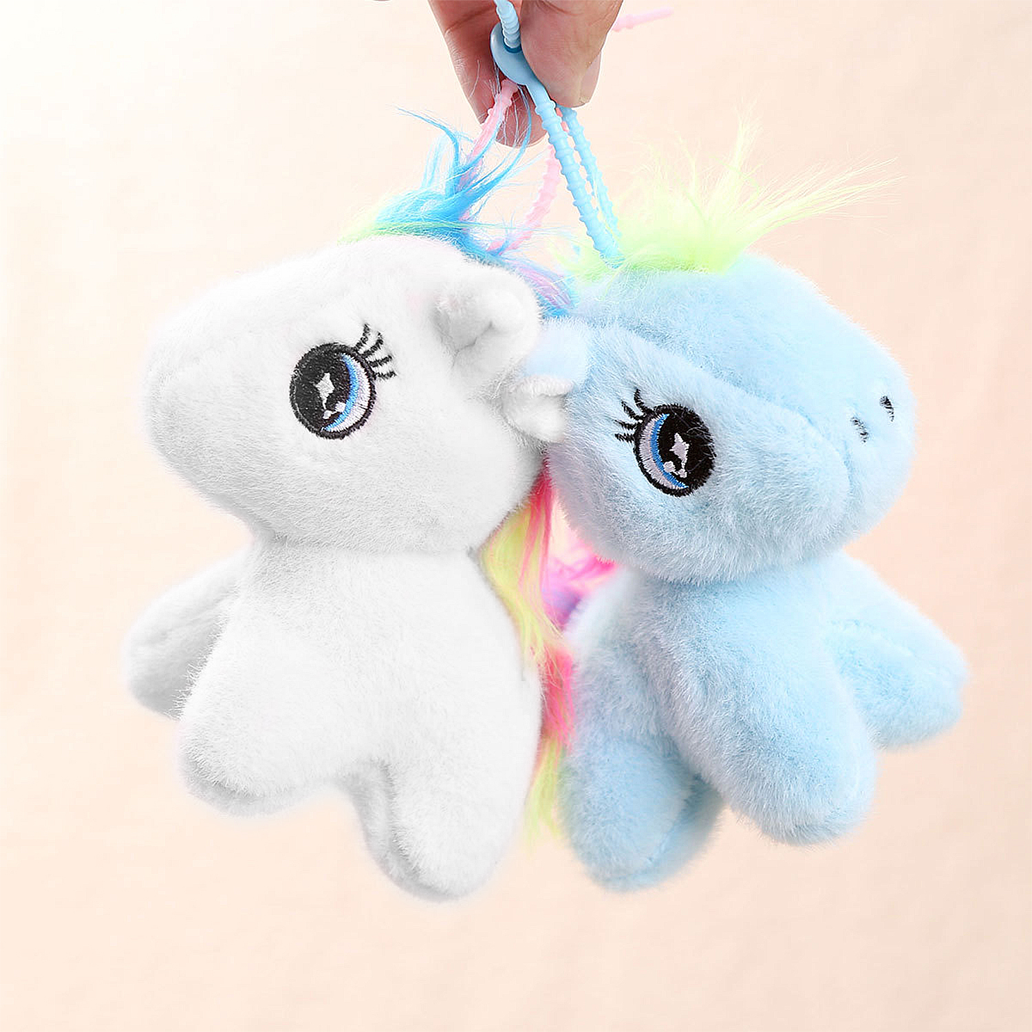 Due portachiavi a forma di unicorno di peluche. Uno bianco, uno blu, entrambi con criniere colorate. Tenuti da una mano su sfondo rosa.