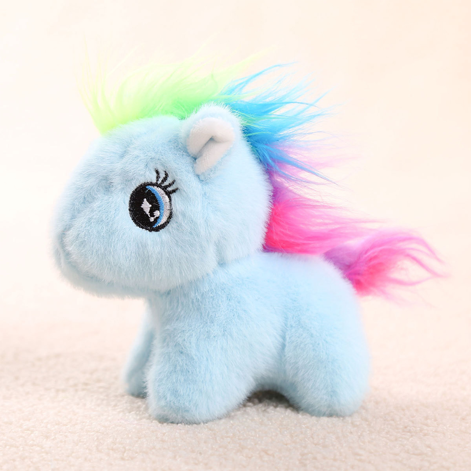 Un pony di peluche blu con criniera e coda arcobaleno, occhi neri e orecchie bianche. Su una superficie morbida e chiara.
