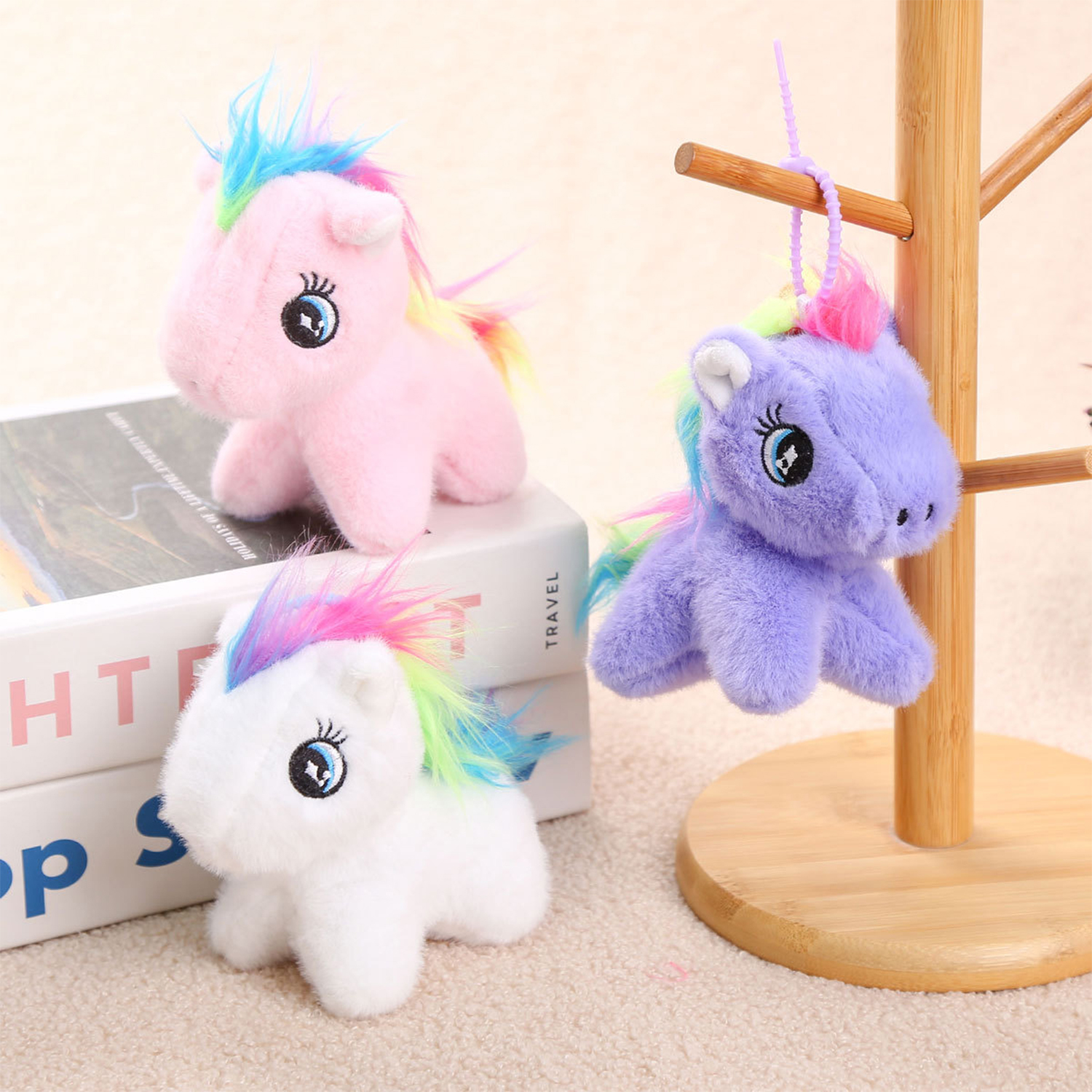 Tre unicorni di peluche con criniere e code arcobaleno. Uno è rosa, uno è viola e uno è bianco. Sono su una superficie chiara.