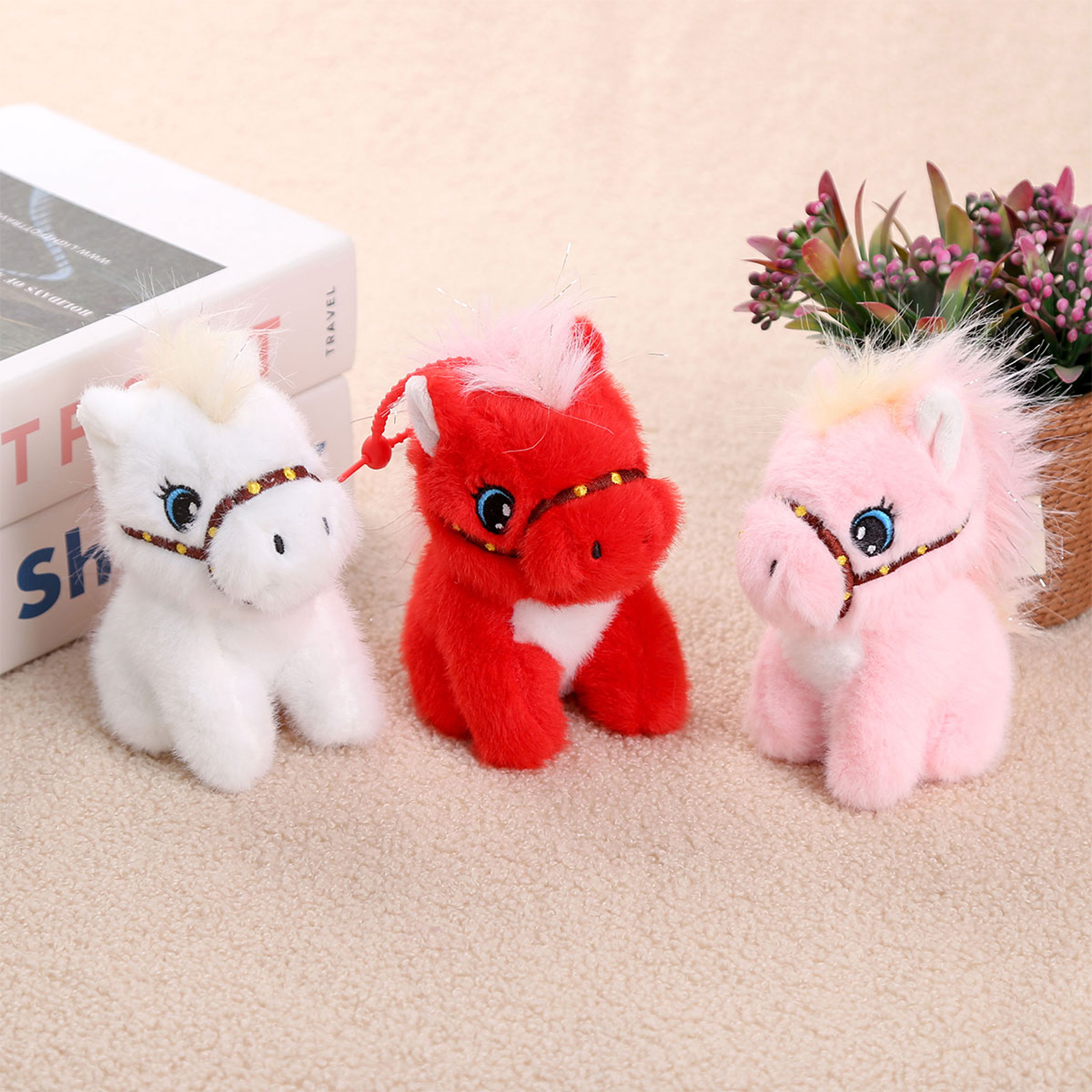 Tre cavalli di peluche: bianco, rosso e rosa. Hanno occhi blu e finimenti marroni. Libri e fiori sullo sfondo.