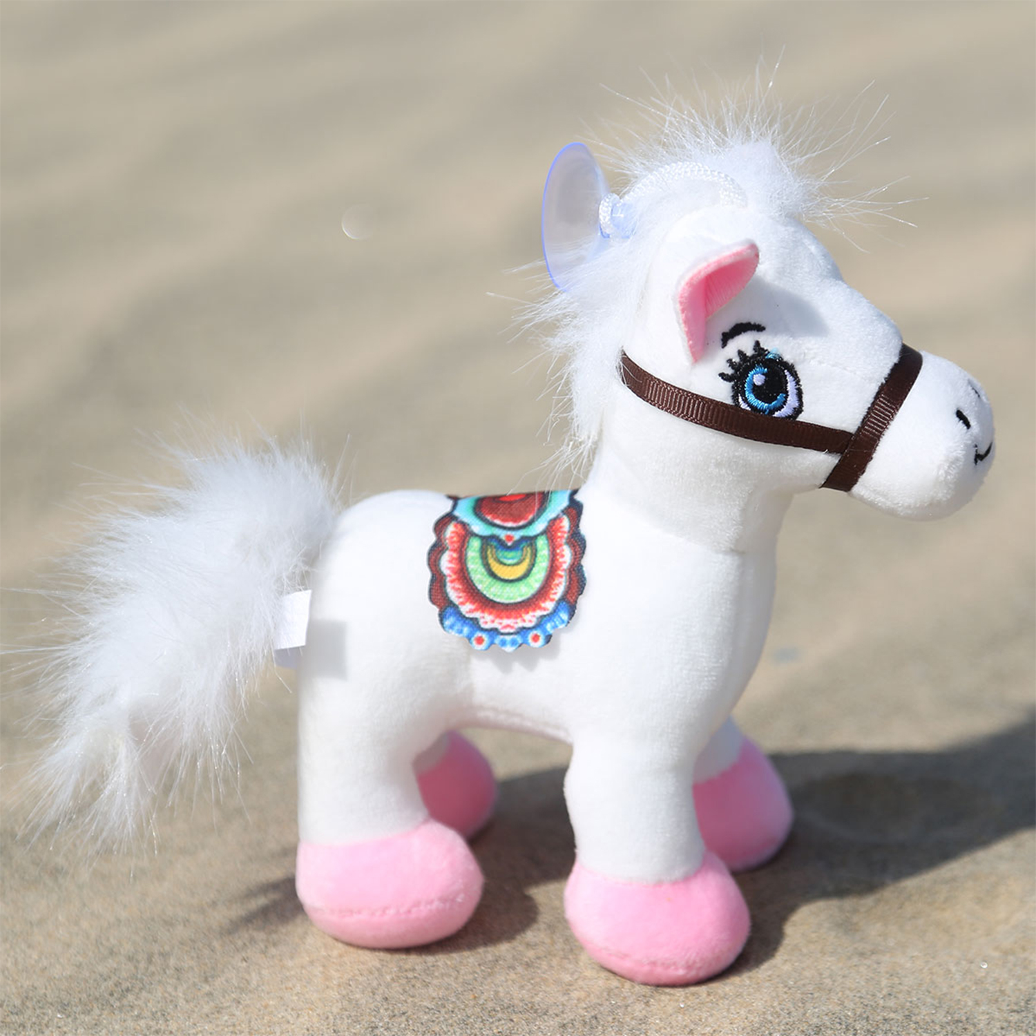 Un cavallo di peluche bianco con zoccoli rosa, una sella colorata e una ventosa sulla testa è sulla sabbia.