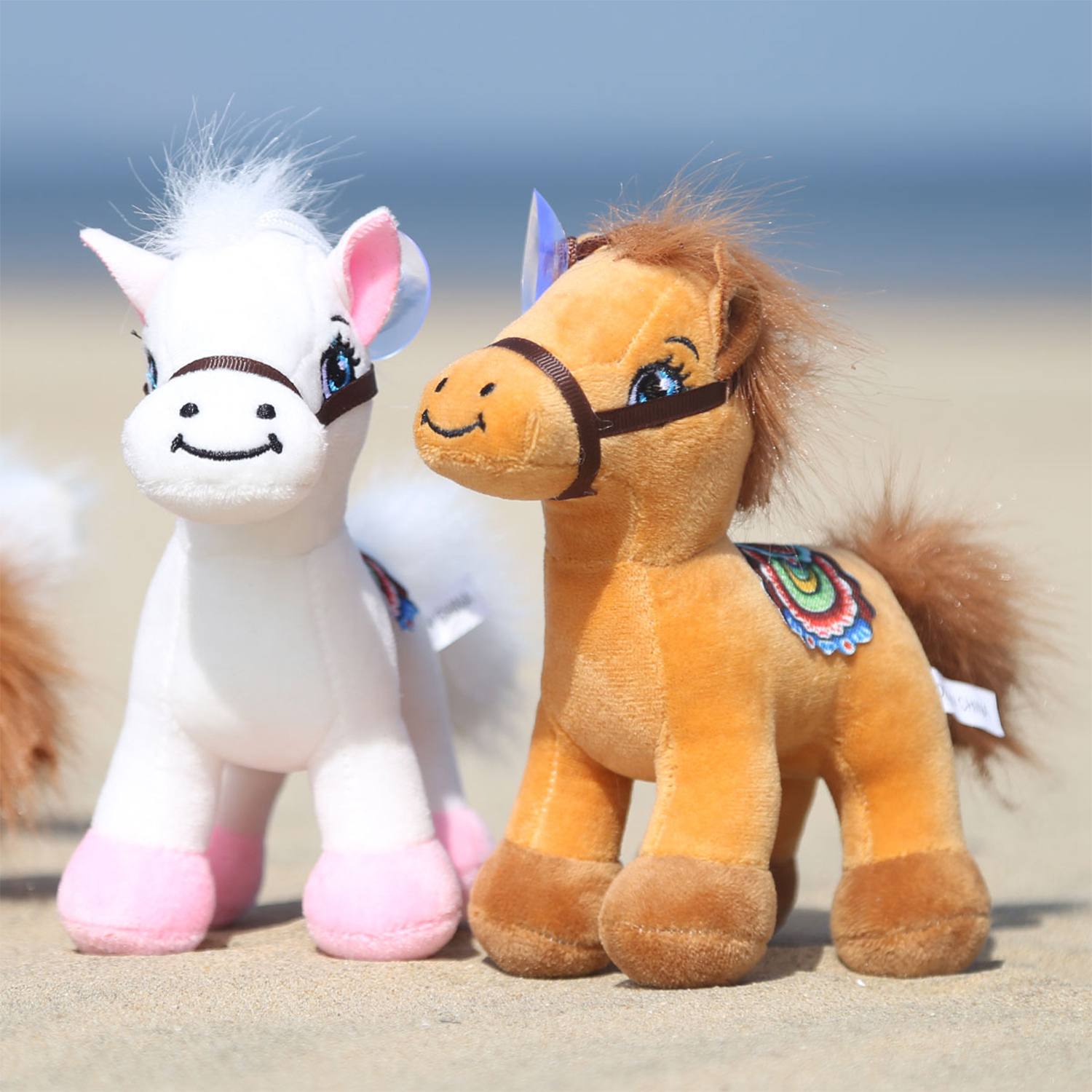 Due cavalli di peluche sulla sabbia. Uno è bianco con dettagli rosa, l'altro è marrone chiaro. Lo sfondo mostra il mare e il cielo.