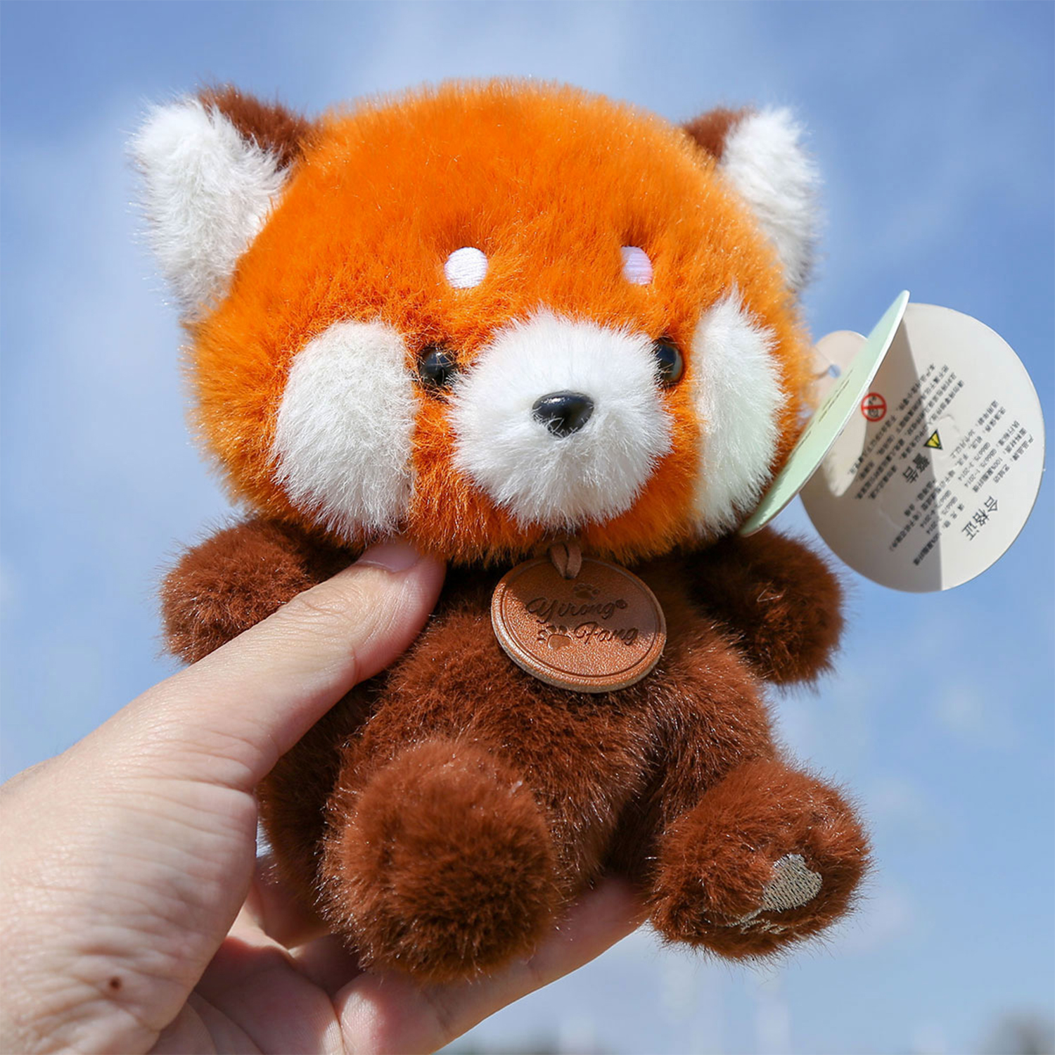 Una mano tiene in mano un peluche di panda rosso. Ha la pelliccia arancione con accenti bianchi e un'etichetta di cuoio marrone. Lo sfondo è un cielo blu.