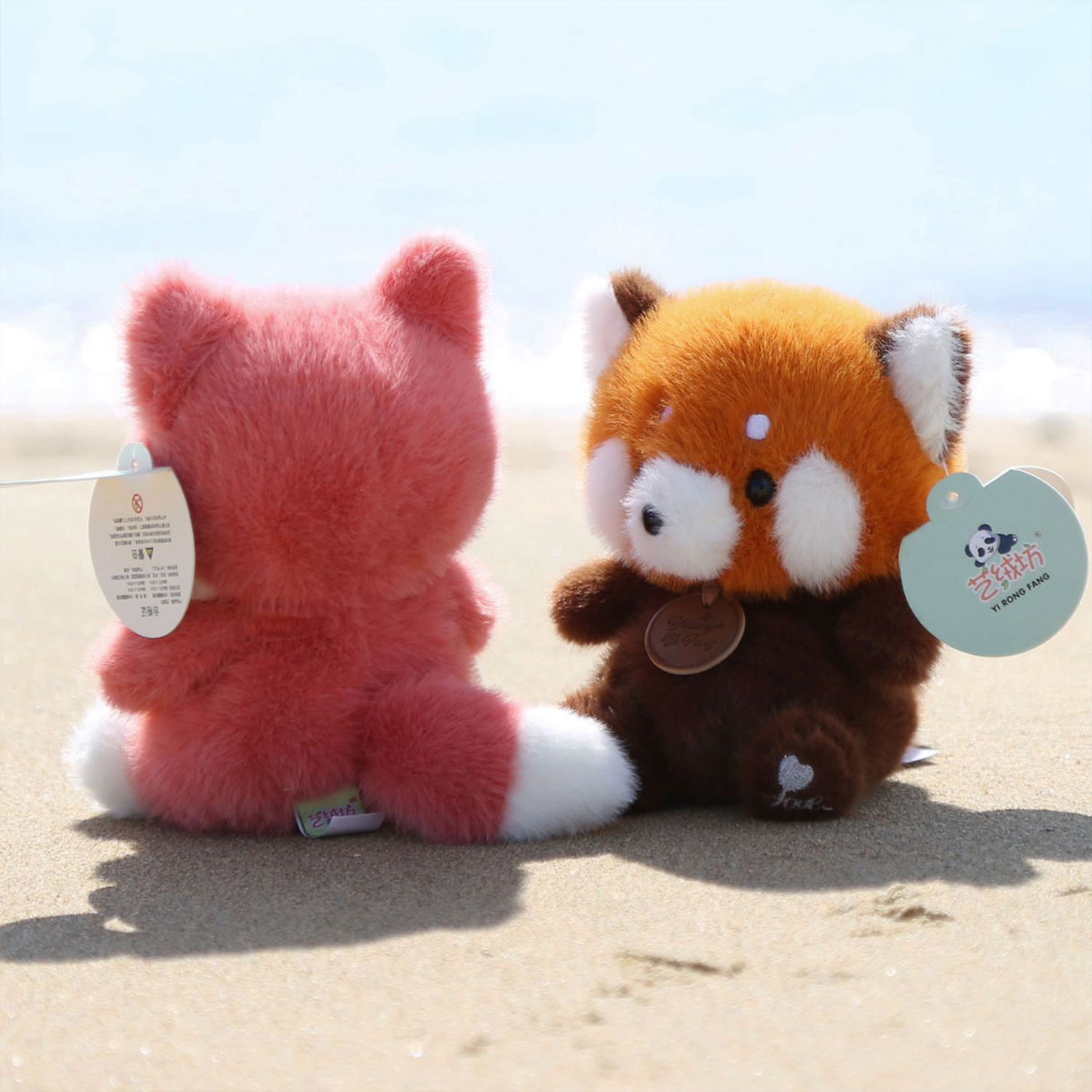 Due panda rossi di peluche sulla sabbia vicino al mare. Uno è rosa, l'altro marrone e arancione. Entrambi hanno etichette.