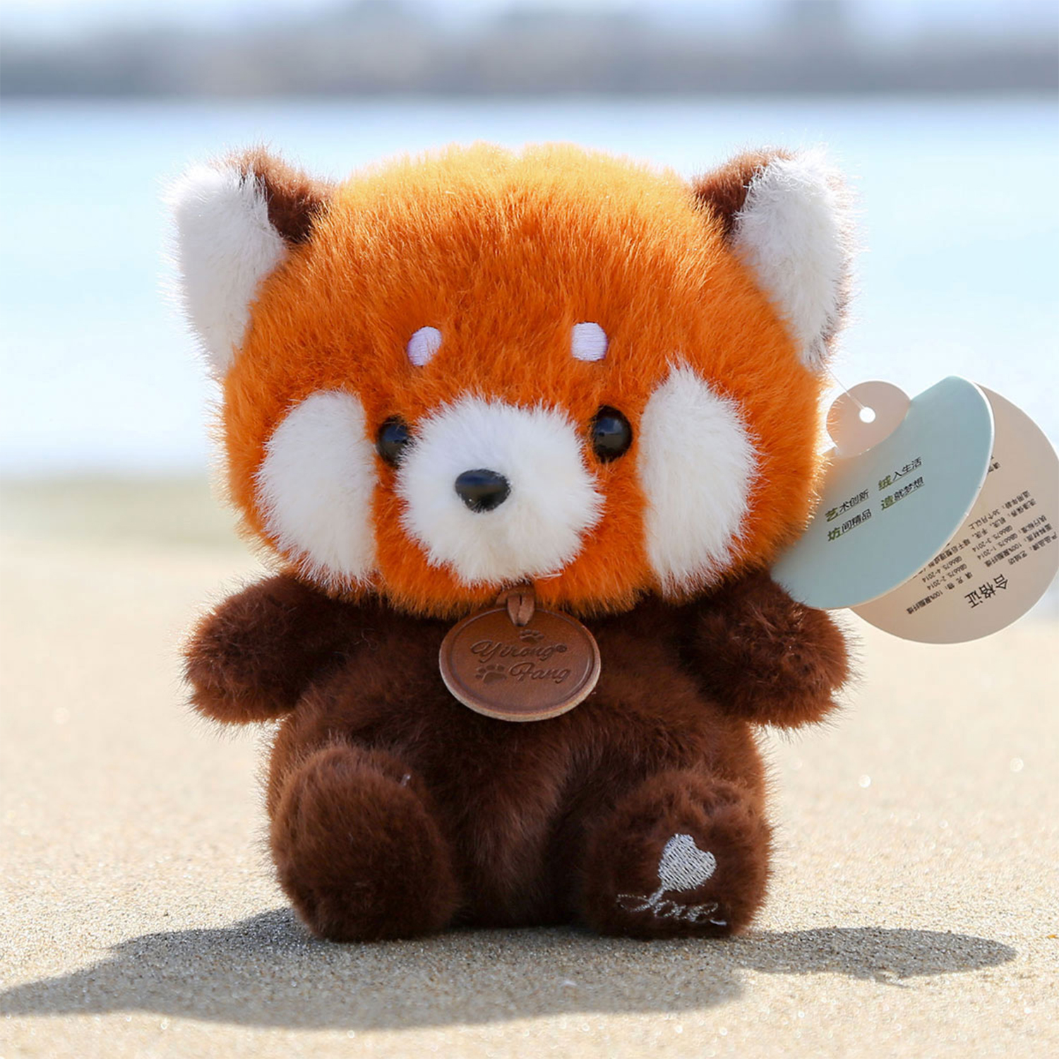 Un peluche di panda rosso con dettagli bianchi, corpo marrone e un'etichetta, seduto sulla sabbia, con l'oceano sfocato sullo sfondo.