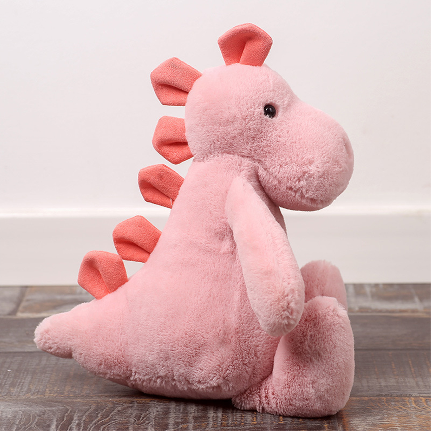 Un giocattolo di dinosauro di peluche rosa con spine e orecchie arancioni è seduto su una superficie di legno contro un muro bianco.