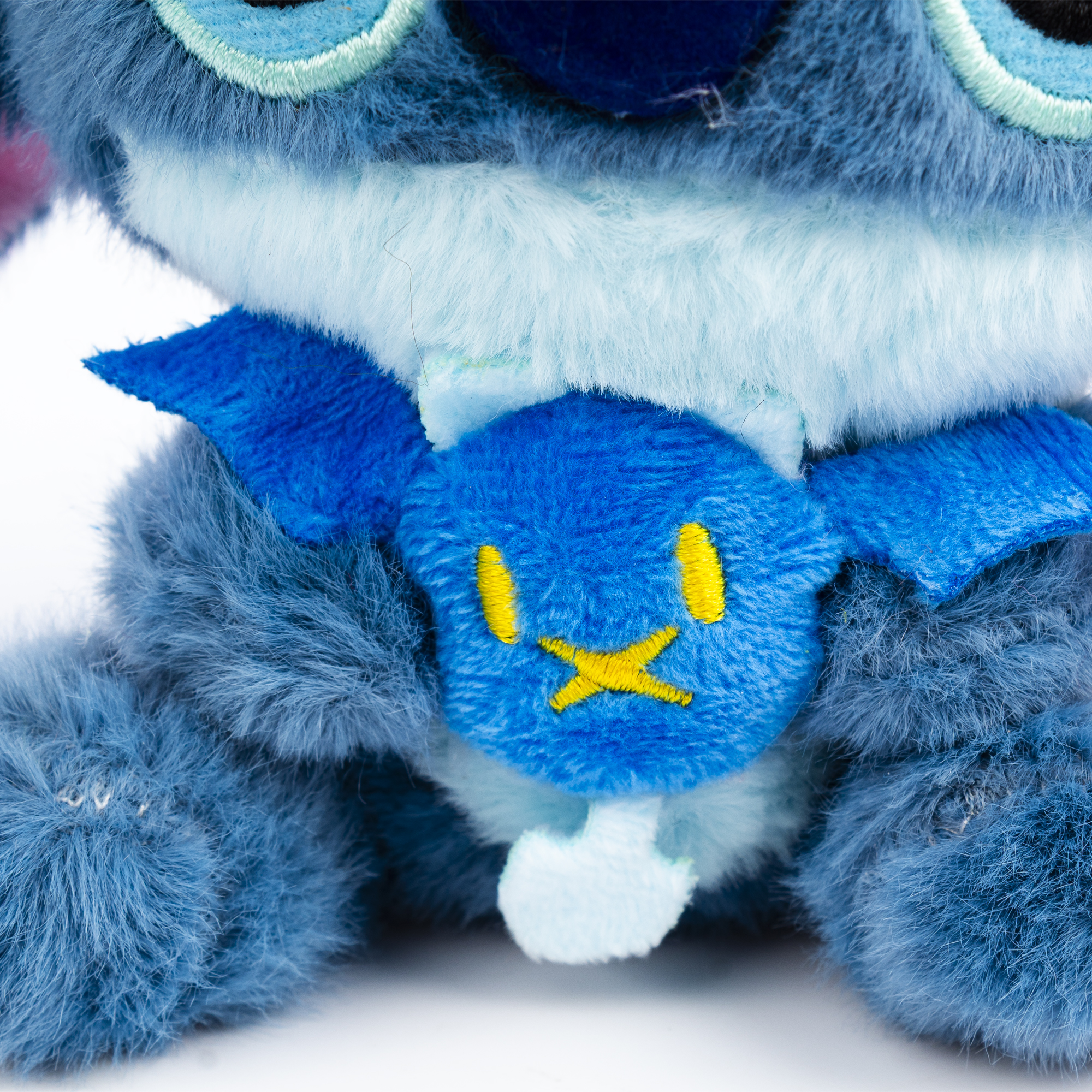 Primo piano di un peluche di Stitch blu con dettagli ricamati gialli, su sfondo bianco.