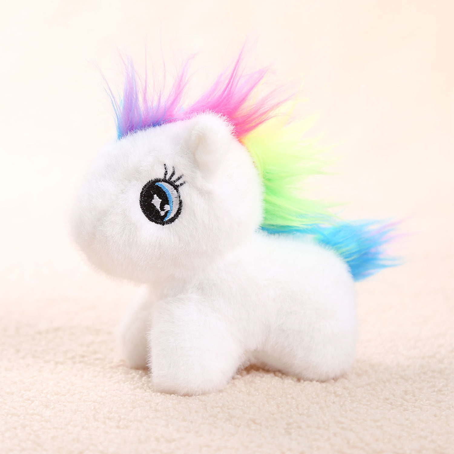 Un peluche di unicorno bianco con criniera e coda arcobaleno. Ha grandi occhi blu ed è seduto su una superficie beige strutturata.