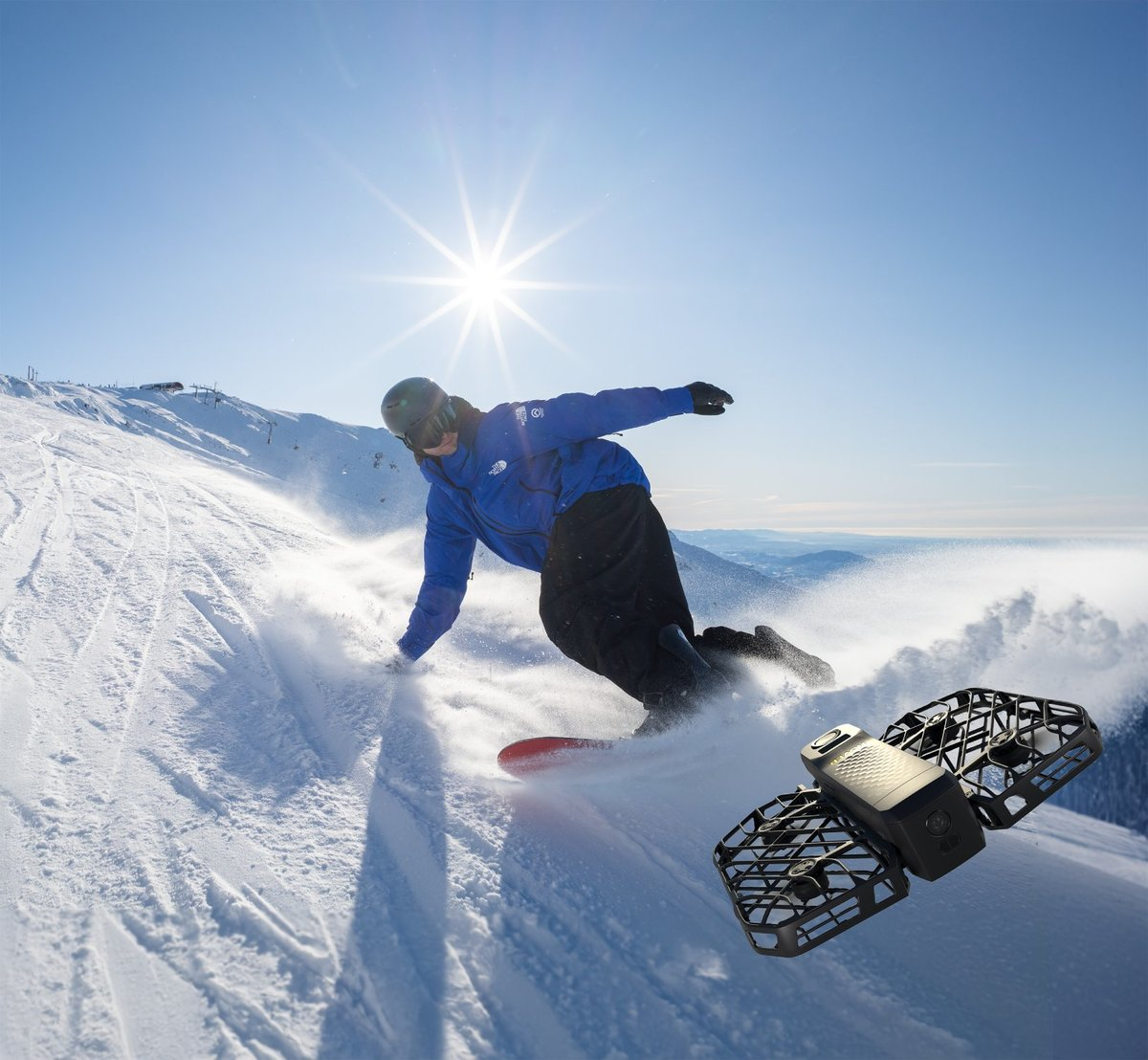 Snowboardzista zjeżdża ze śnieżnego stoku w jasnym słońcu. Dron unosi się w pobliżu. W tle góry i błękitne niebo.