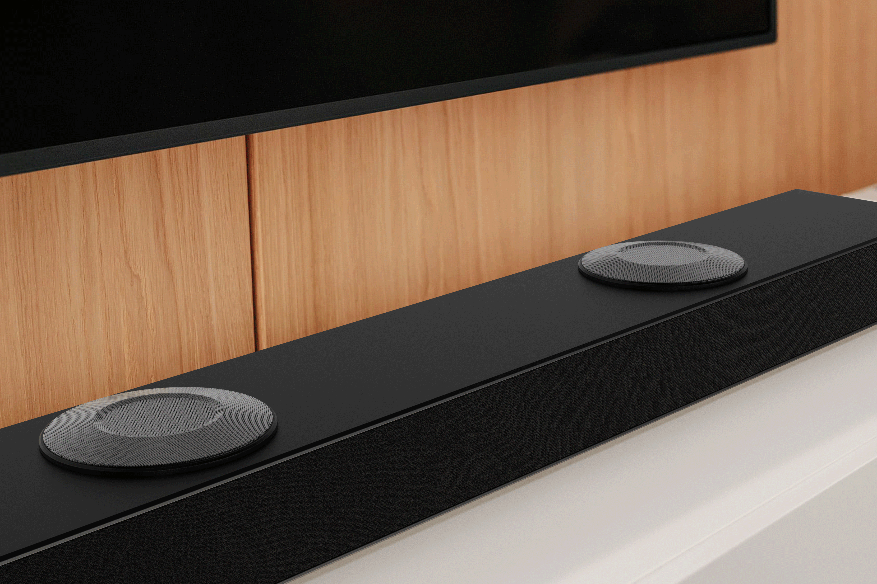 Una barra de sonido negra con dos elementos circulares grises. Paneles de madera y TV negro al fondo.