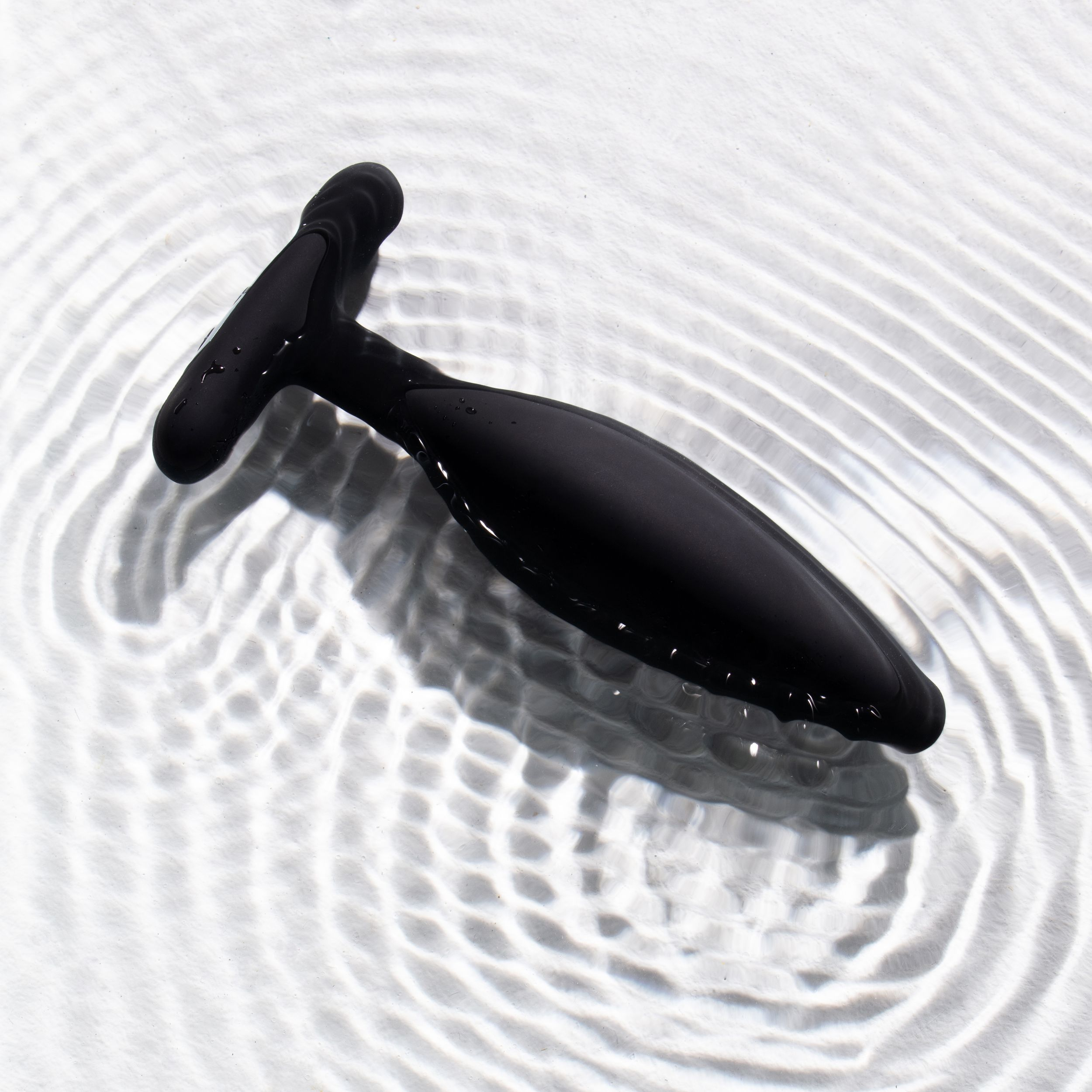 Ein schwarzes Sexspielzeug liegt im Wasser und reflektiert die Wellen. Das Spielzeug hat einen Griff und einen glatten, sich verjüngenden Körper.