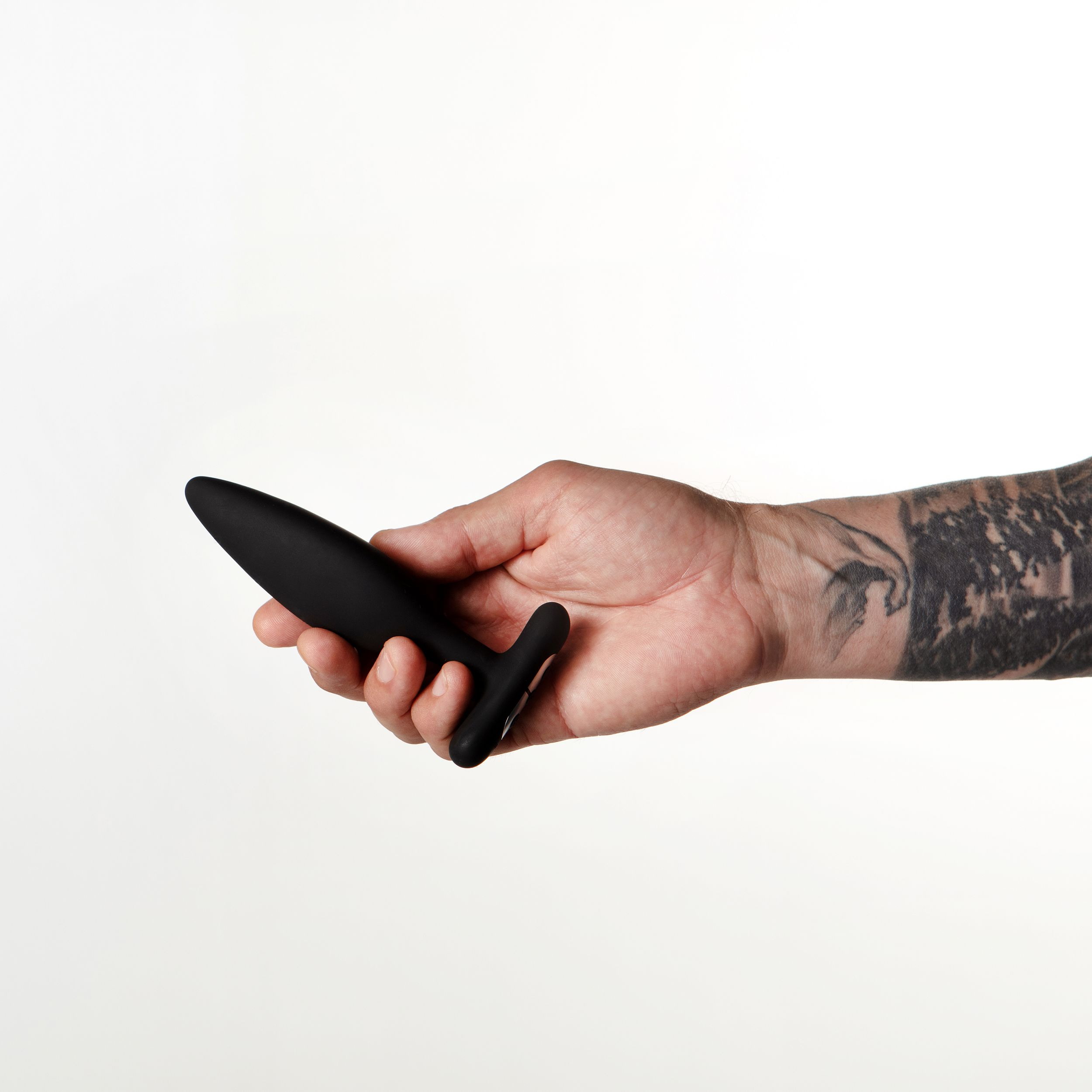 Eine Hand hält ein schwarzes Sexspielzeug vor weißem Hintergrund. Der Arm hat ein Tattoo.