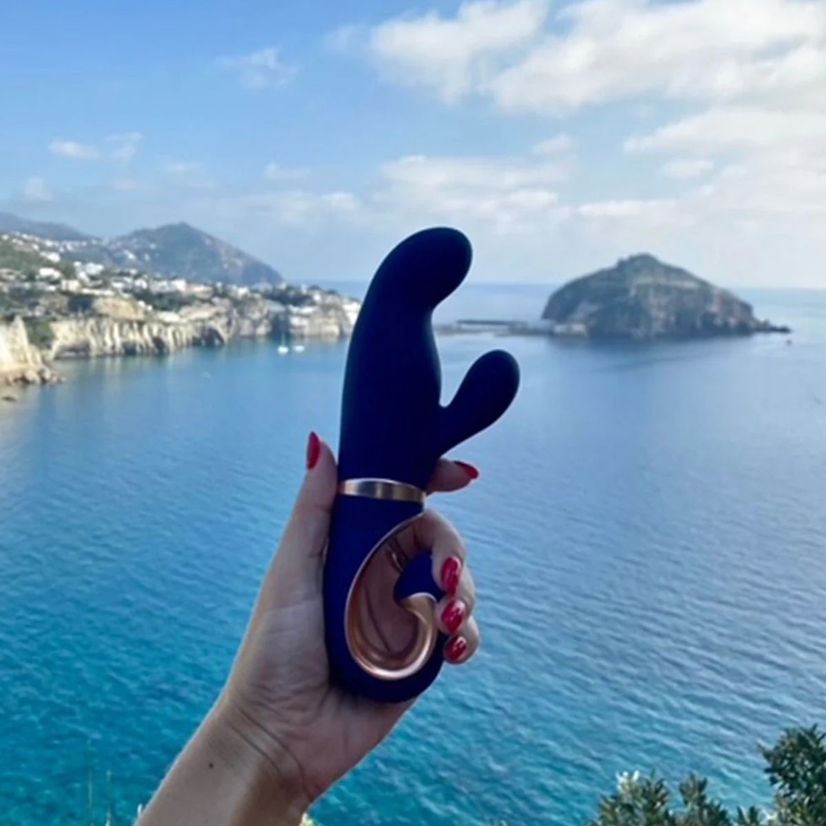 Eine Hand hält einen blauen Vibrator mit goldenem Ring, mit Blick auf das Meer mit Inseln und Bergen im Hintergrund.
