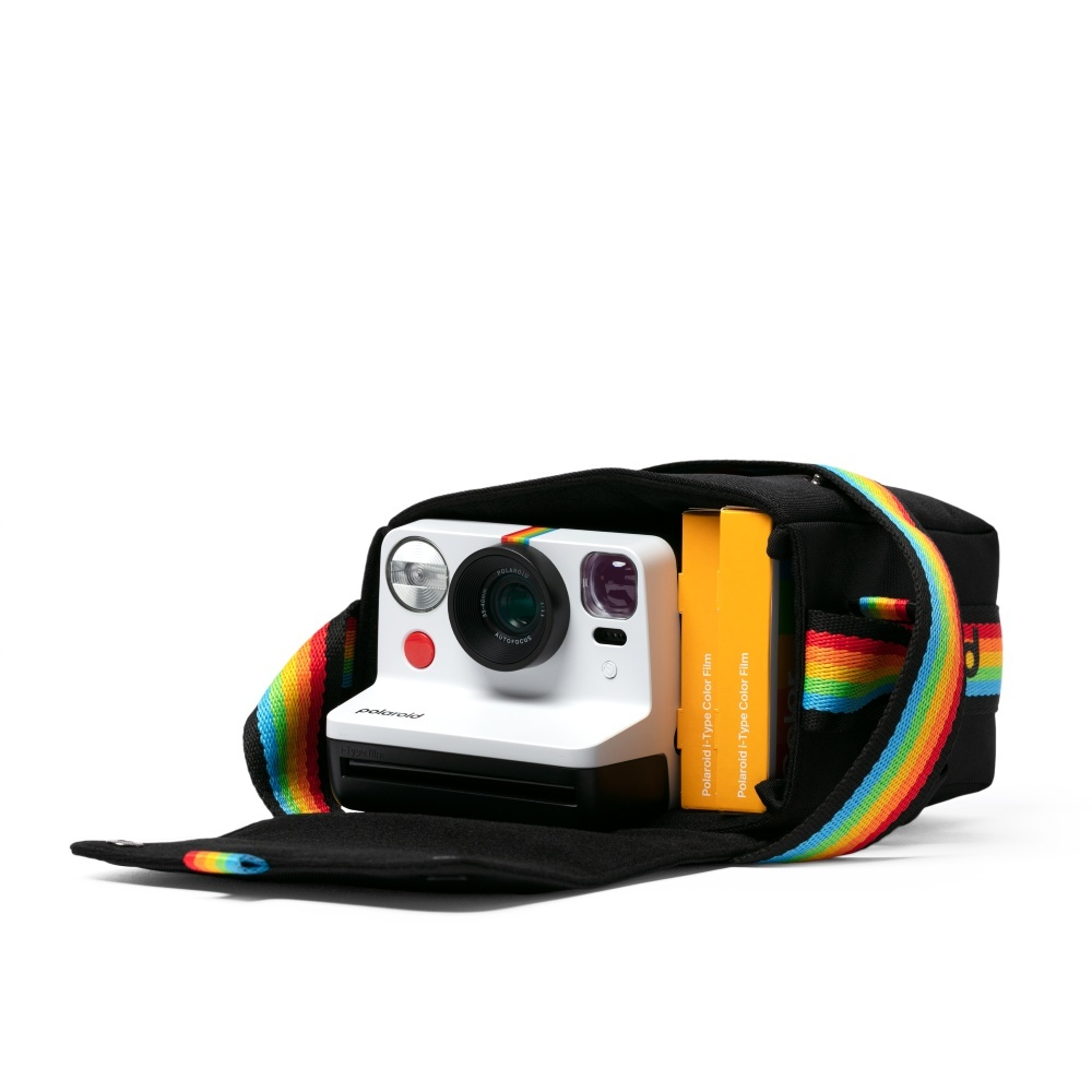 Eine weiße Polaroid-Kamera befindet sich in einer schwarzen Tasche mit Regenbogenriemen. Zwei gelbe Filmboxen sind ebenfalls vorhanden. Der Hintergrund ist weiß.