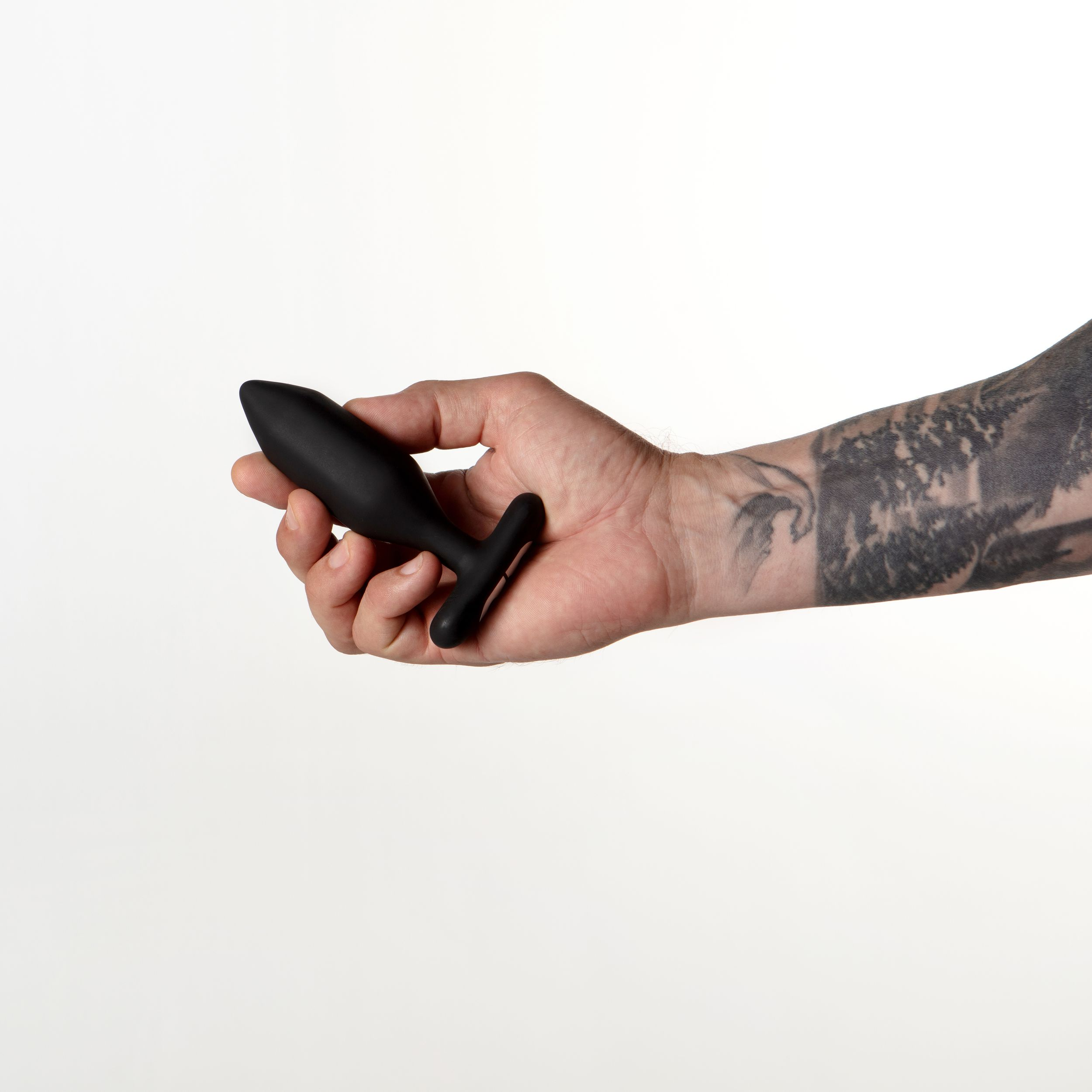 Eine Hand hält einen schwarzen Analplug vor weißem Hintergrund. Der Arm hat ein Tattoo.