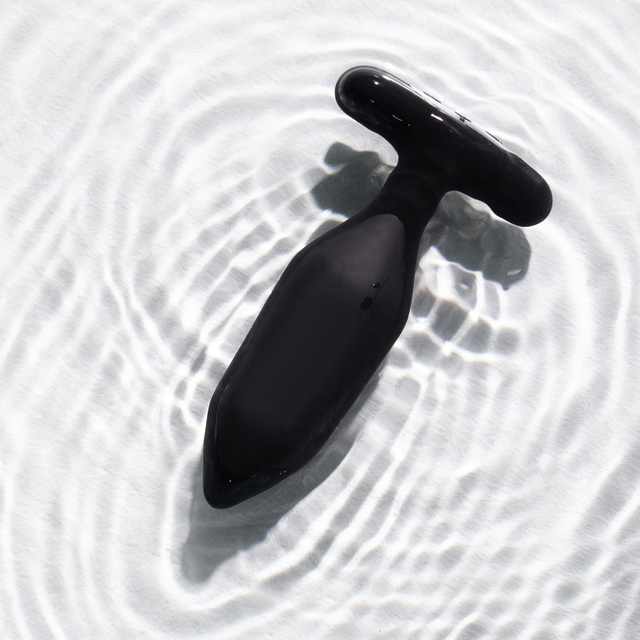 Ein schwarzes Sexspielzeug liegt auf welligem Wasser. Es hat einen langen, gebogenen Körper und einen T-förmigen Griff.