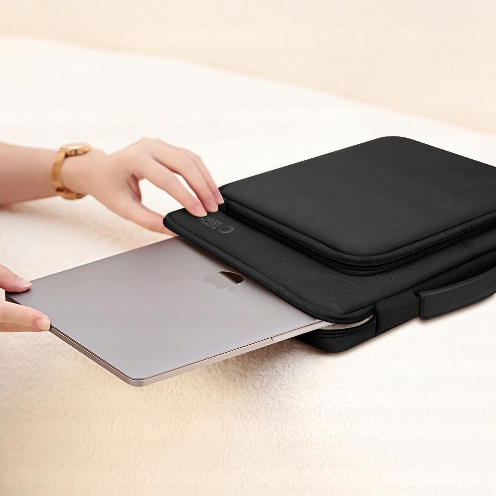 Een persoon stopt een zilveren laptop in een zwarte laptoptas. De tas ligt op een lichtgekleurd oppervlak.