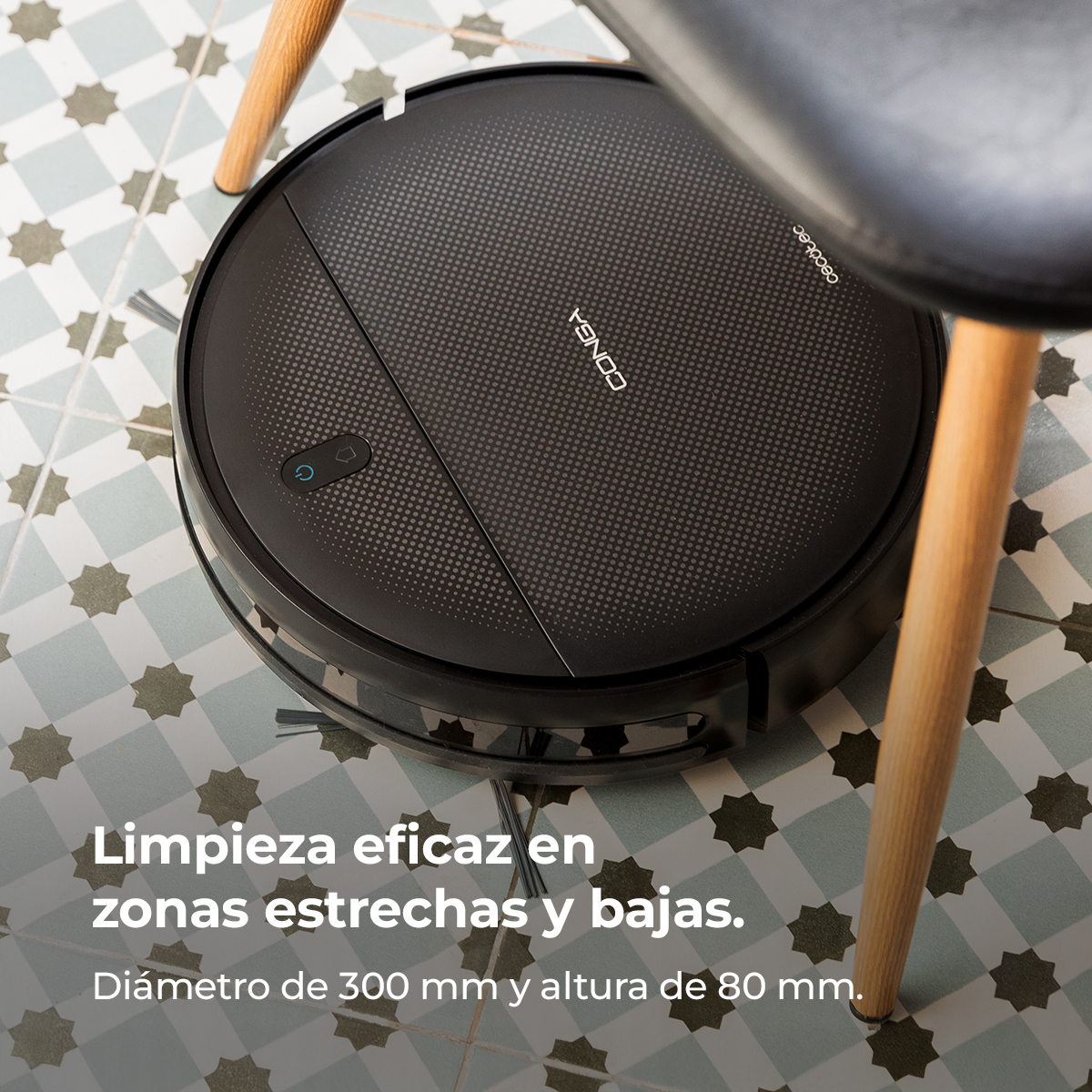 Una aspiradora robot negra debajo de una silla. La aspiradora está en un suelo de baldosas estampadas. El texto dice limpieza eficiente en áreas estrechas.