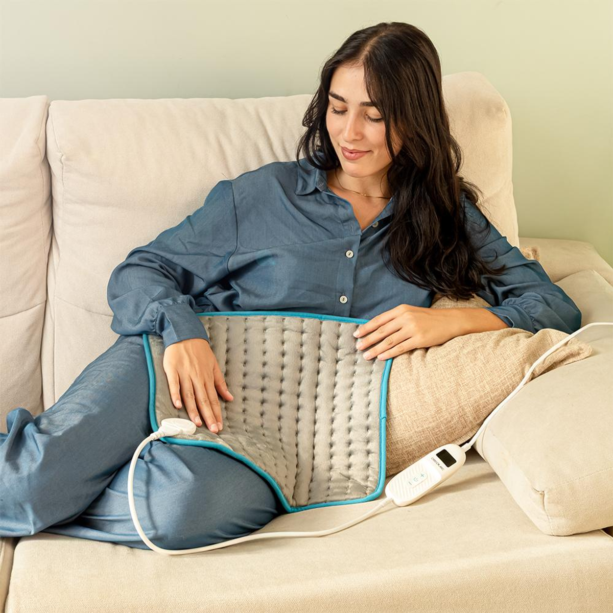 Una mujer con ropa azul se sienta en un sofá. Usa una almohadilla térmica gris. Un mando a distancia está en el sofá.