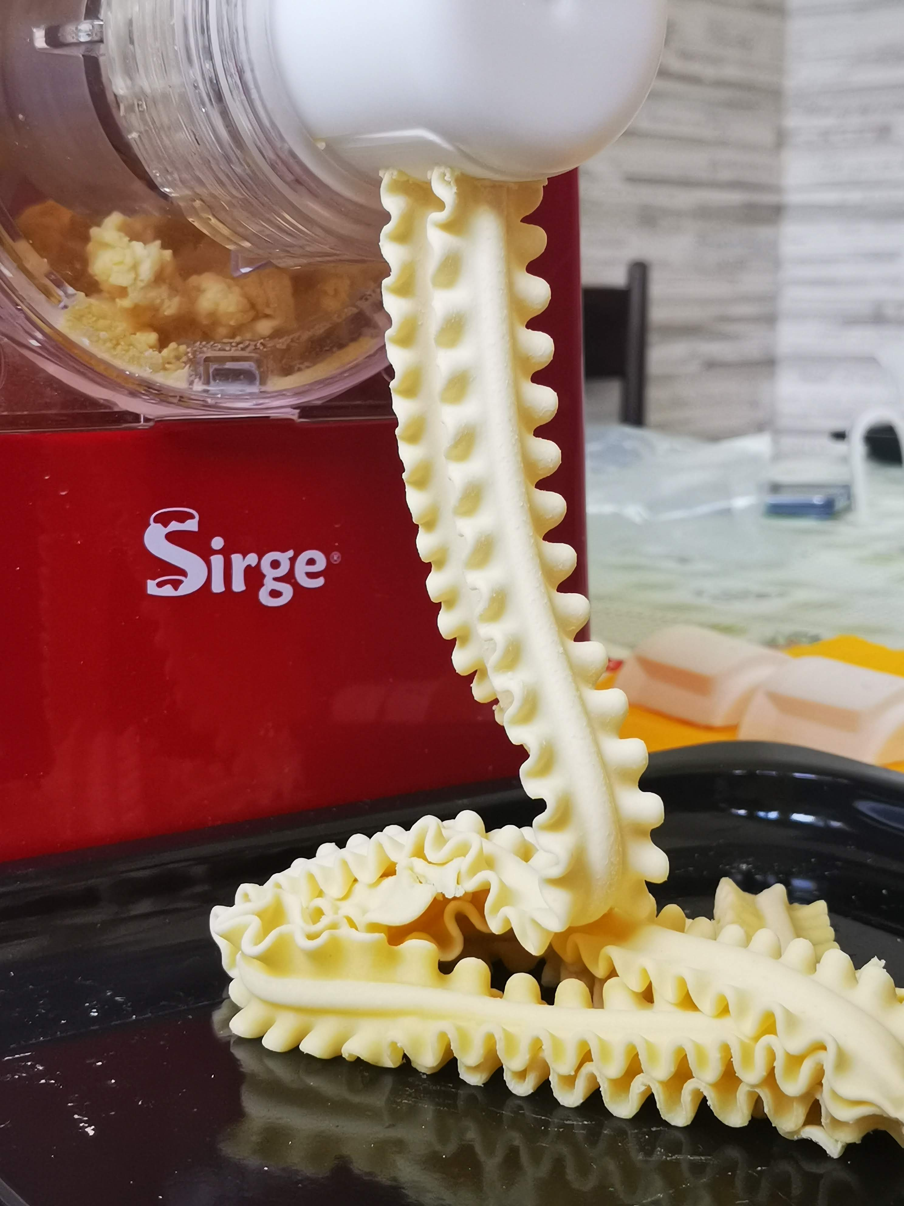 Una macchina per la pasta estrude tagliatelle di lasagne fresche e arruffate. Macchina rossa e vassoio nero in primo piano. Pasta bianca.