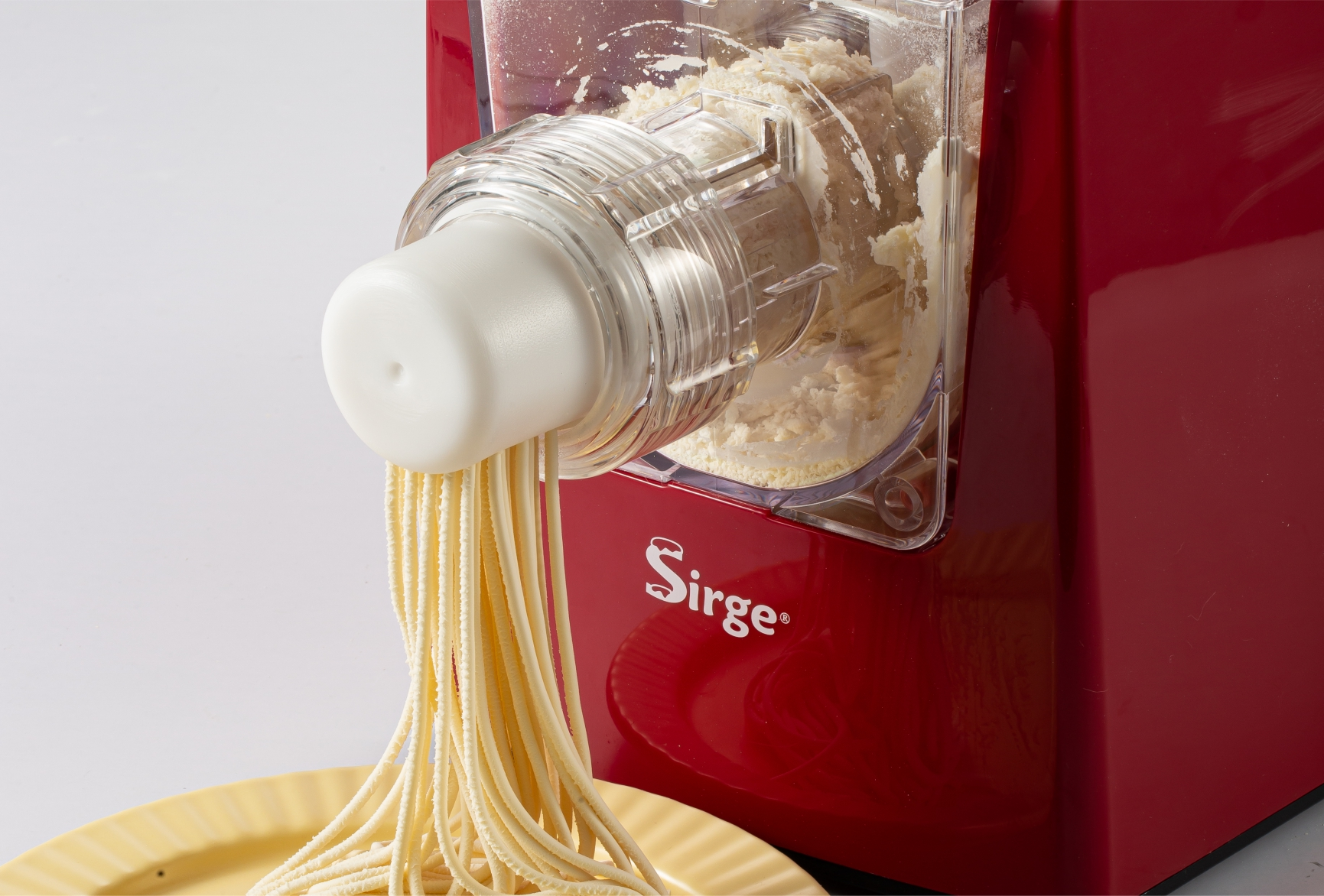 Una macchina per la pasta rossa estrude spaghetti freschi su un piatto giallo. La macchina ha un contenitore trasparente con farina.