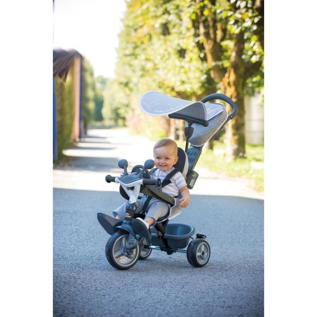 Ein Baby sitzt in einem Dreirad auf einer asphaltierten Straße. Das Dreirad ist dunkelblau mit Sonnenschutz. Das Baby lächelt.