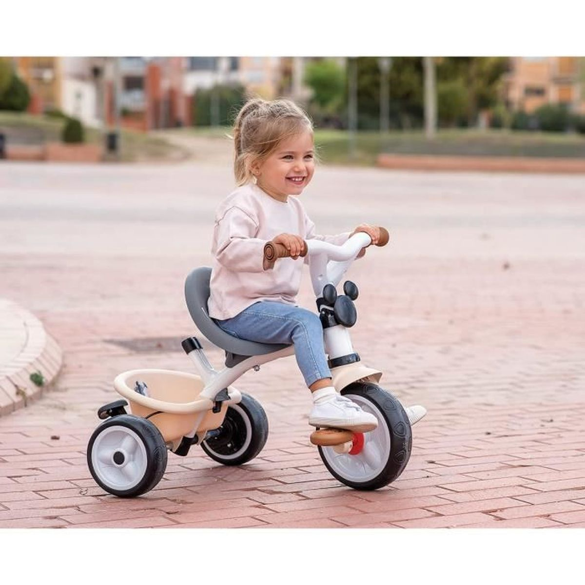 Ein lächelndes Kleinkind fährt auf einem Dreirad auf einem Ziegelweg. Das Dreirad ist beige und weiß. Das Kind trägt ein rosa Hemd und blaue Jeans.