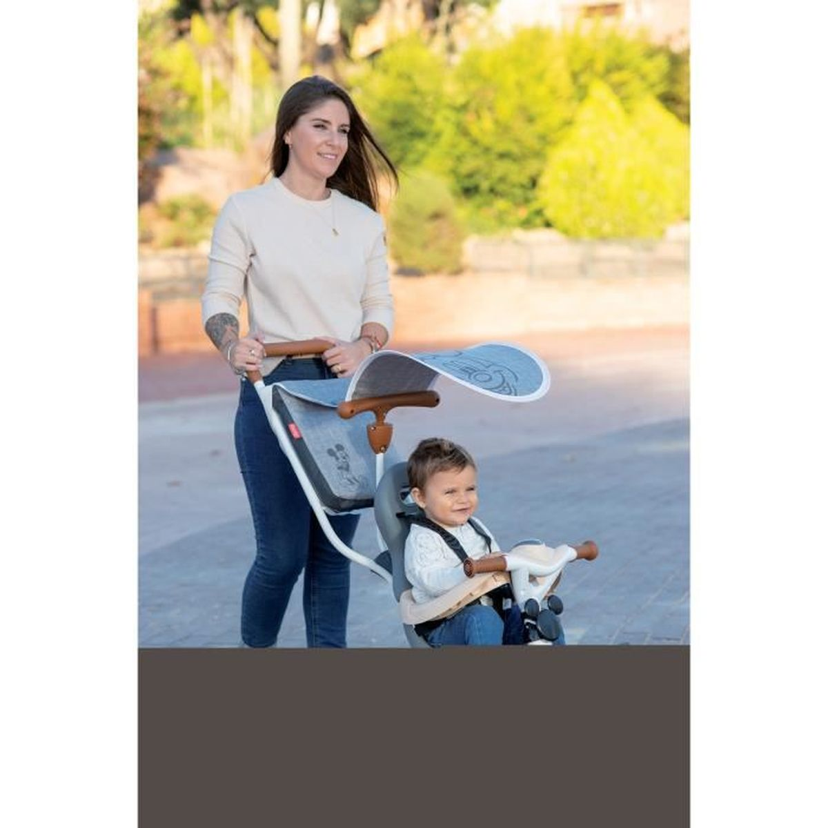 Eine Frau schiebt einen Kinderwagen mit einem Kind. Der Kinderwagen hat ein Verdeck. Sie sind draußen auf einem gepflasterten Bereich.