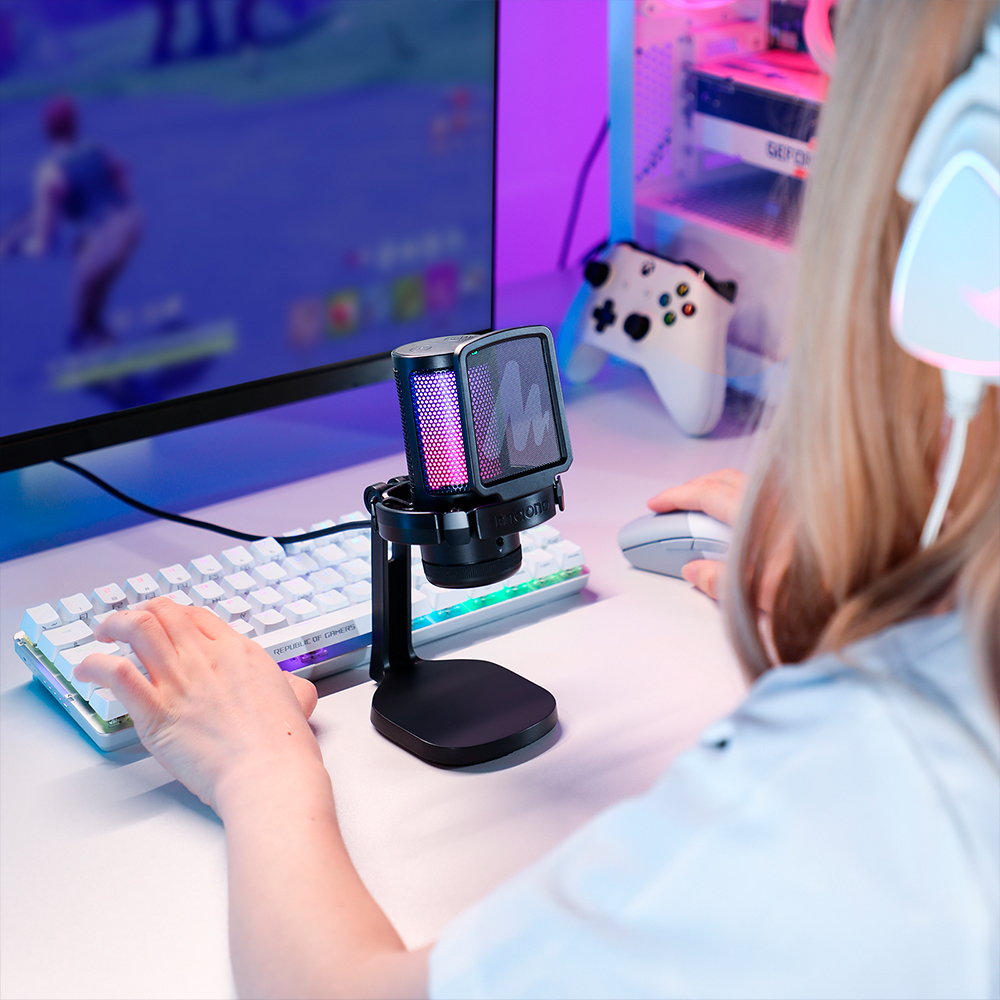 Une personne joue, utilisant un microphone, un clavier et une souris. Un moniteur affiche un jeu. D'autres éléments sont visibles en arrière-plan.