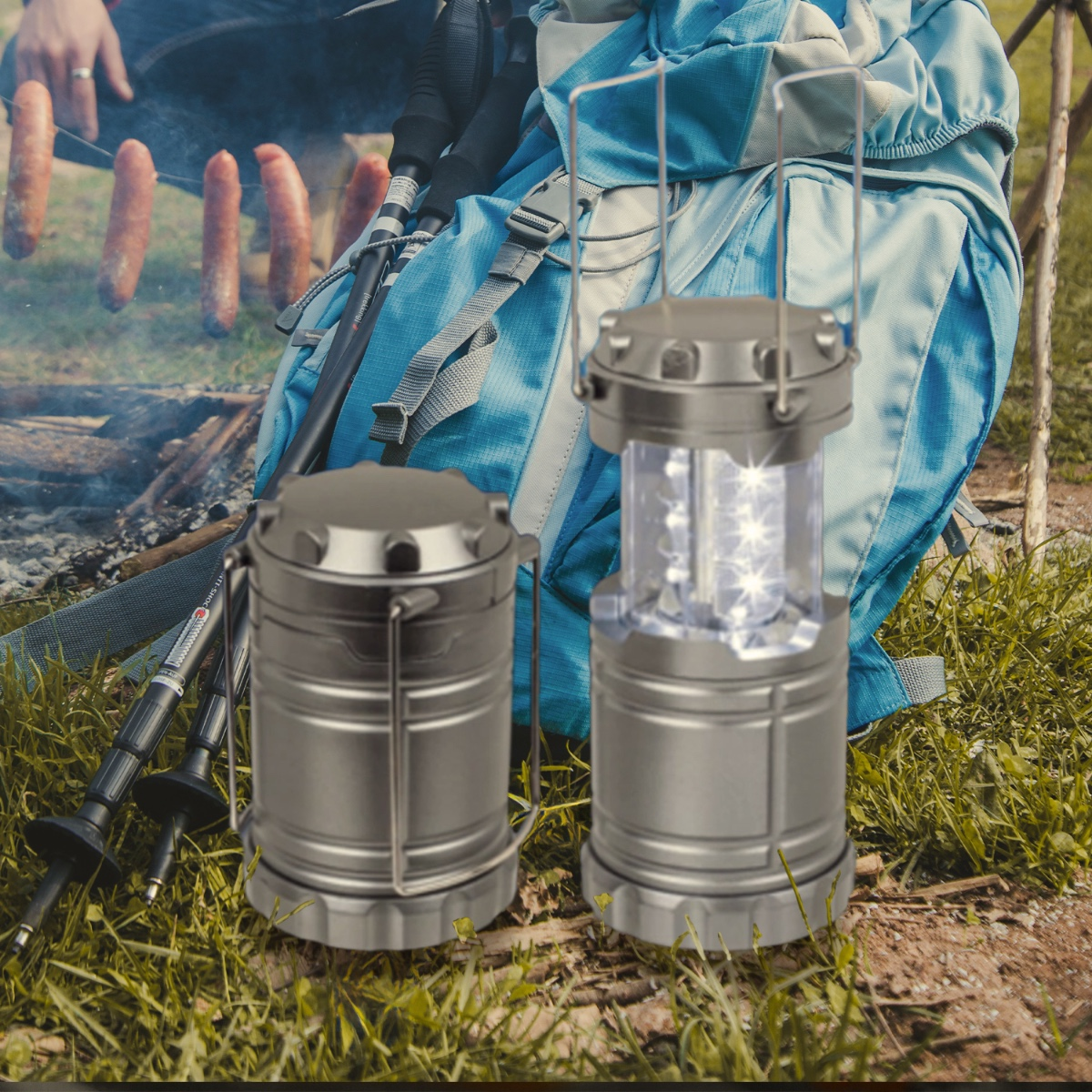 Two metallic camping lanterns sit on grass, with a backpack, trekking poles, and sausages grilling in the background.