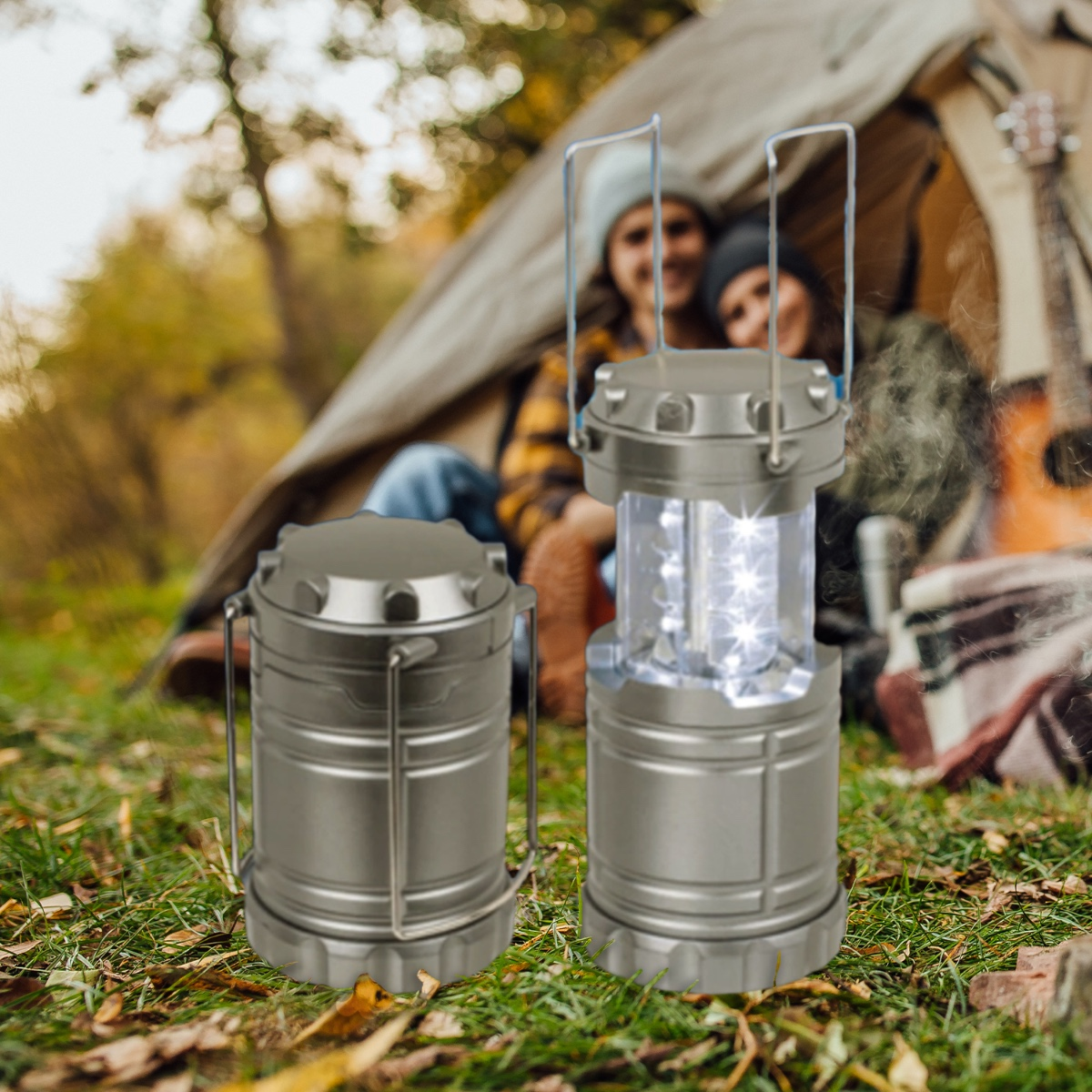 Two silver camping lanterns on green grass, with a tent and blurred people in the background. One lantern is lit.