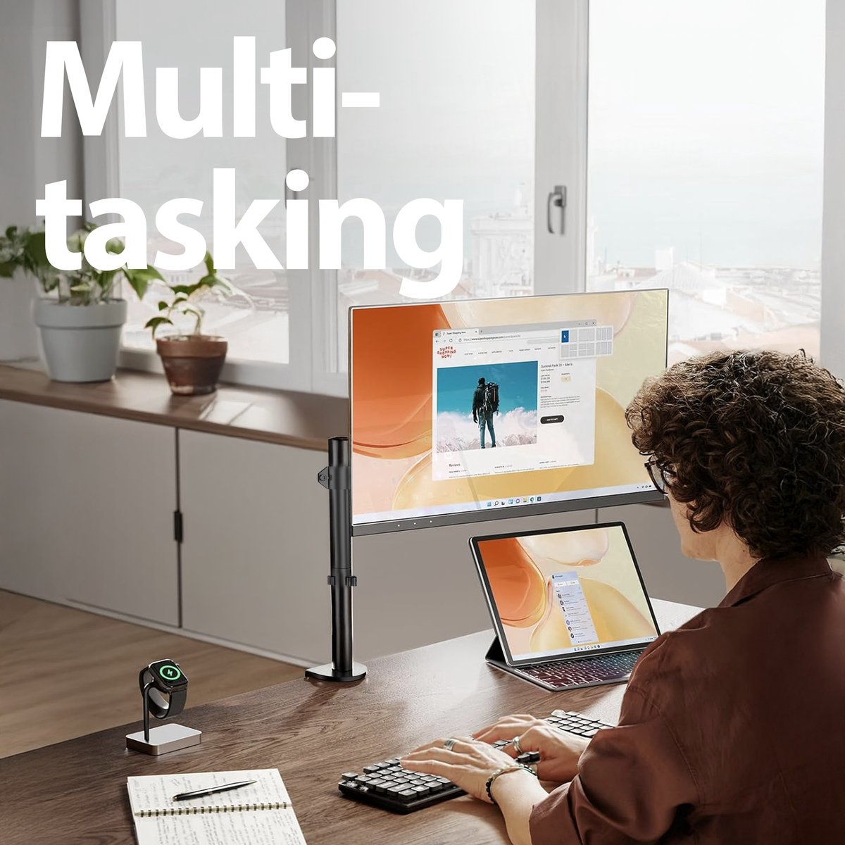 A person is multitasking at a desk with a large monitor and a laptop. There's a smartwatch and keyboard. The background has a window and plants.