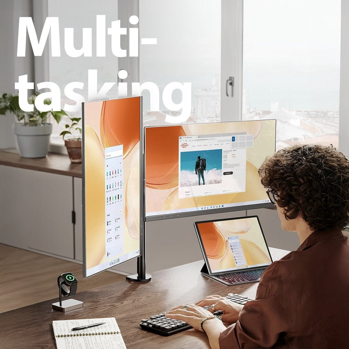 A person is multitasking with multiple screens, a laptop, and a keyboard on a wooden desk. A smartwatch sits on a stand.