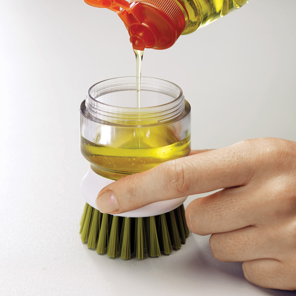 Una mano vierte líquido amarillo de una botella en un recipiente transparente conectado a un cepillo verde, sobre una superficie blanca.