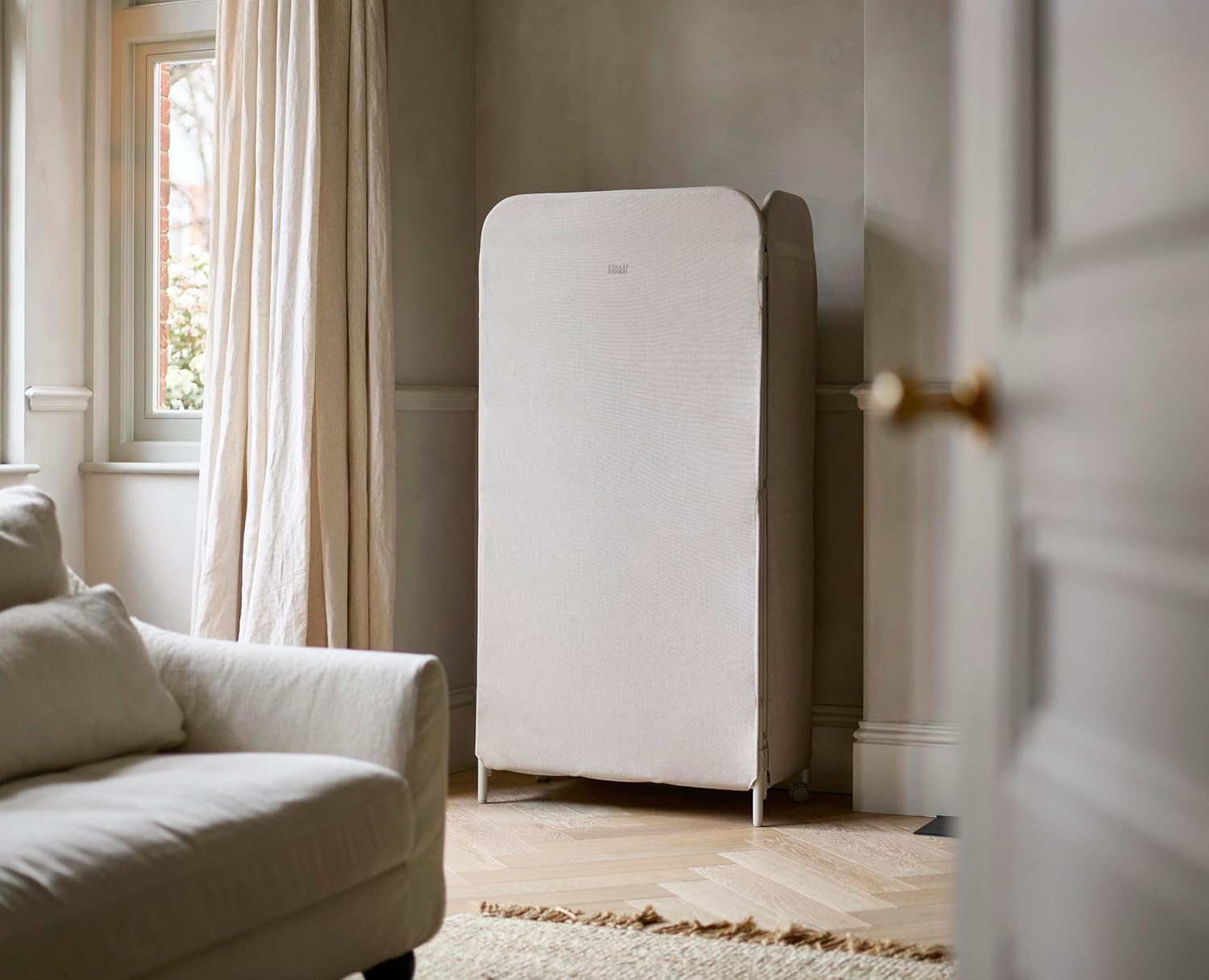 Un purificatore d'aria beige è in una stanza. Sono visibili anche un divano bianco, una finestra con tende e una porta.
