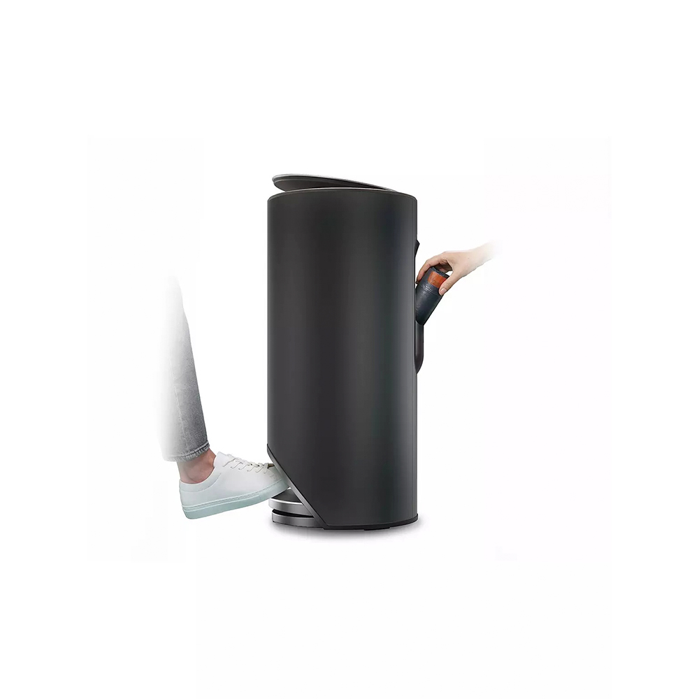 Una persona usa un pedal para abrir un cubo de basura negro. Una mano deja caer algo en el cubo. Se ven zapatos blancos.
