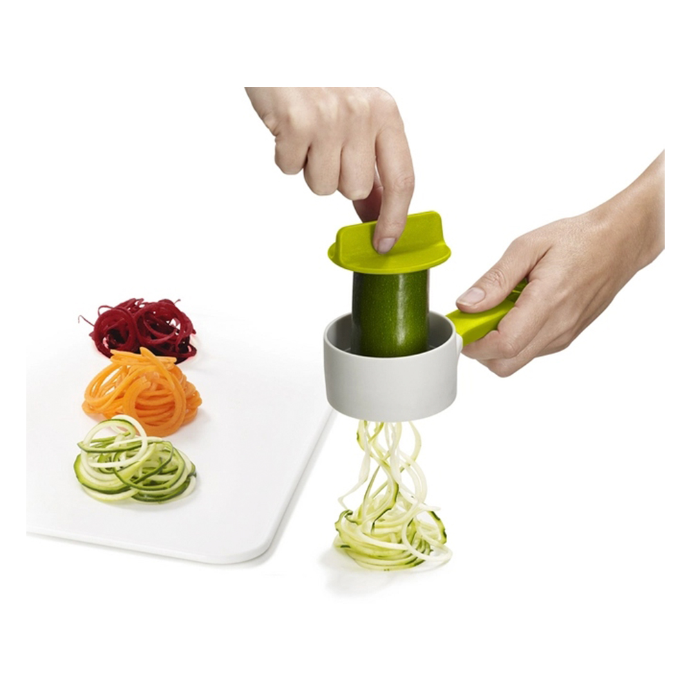 Una persona usa uno spiralizzatore per tagliare zucchine in spaghetti. Tre mucchi di spaghetti di verdure su un tagliere bianco.
