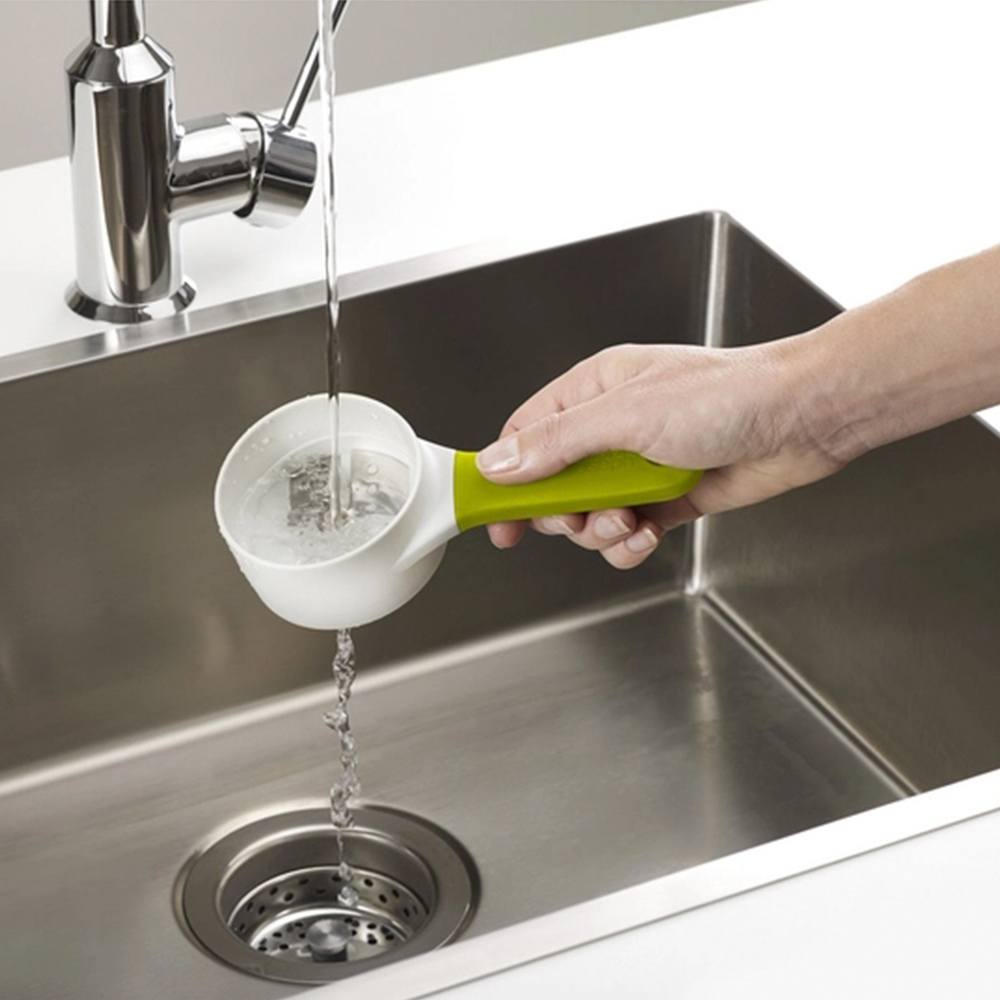 Una mano tiene un misurino bianco e verde sotto un rubinetto, l'acqua vi scorre dentro e trabocca in un lavandino in acciaio inossidabile.