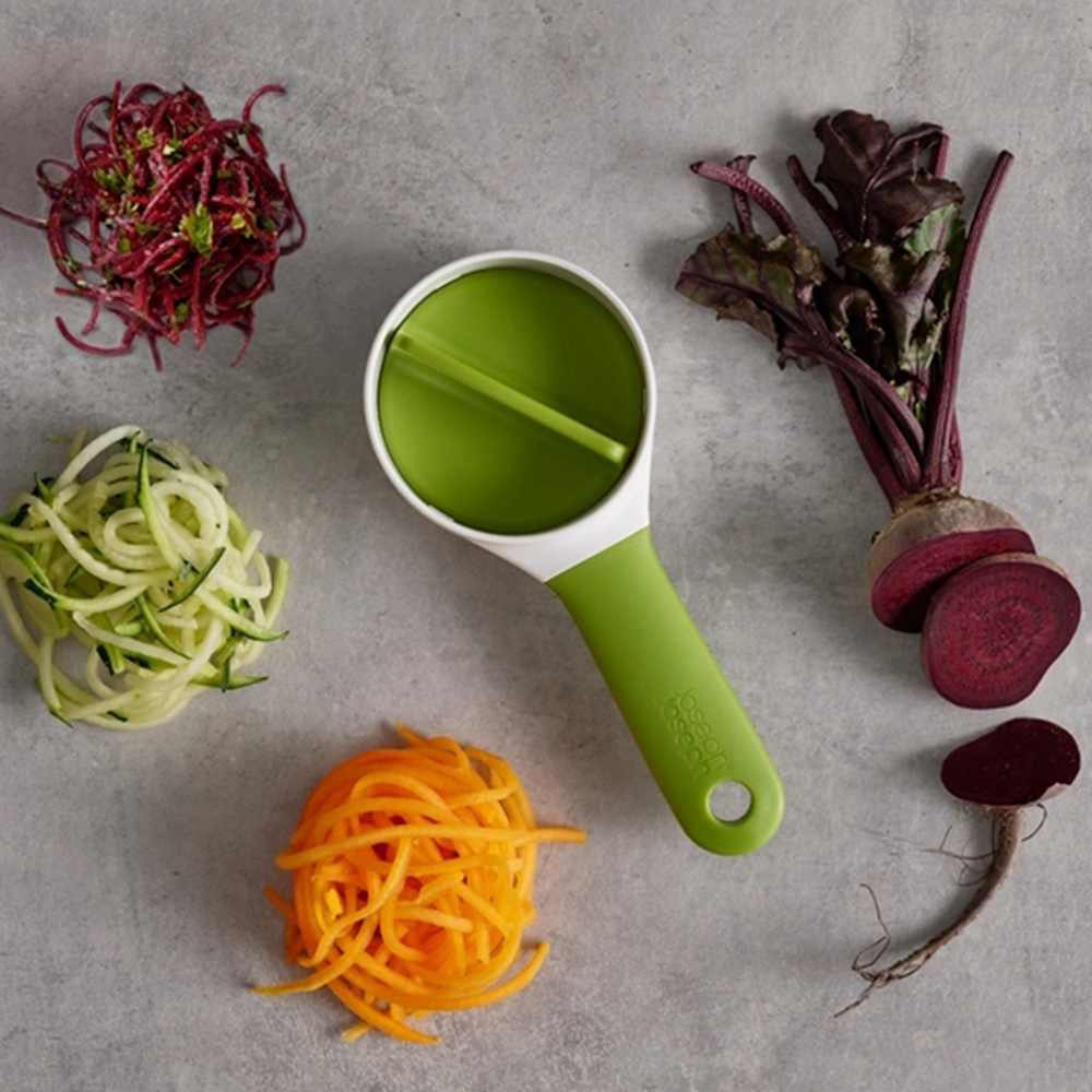 Uno spiralizzatore verde e bianco con spaghetti di verdure, barbabietole e carote su una superficie grigia.