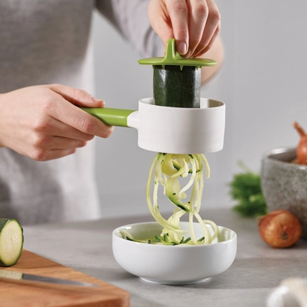 Una persona taglia a spirale zucchine con uno strumento verde e bianco. Gli spaghetti di zucchine cadono in una ciotola bianca. Cipolle e prezzemolo sullo sfondo.