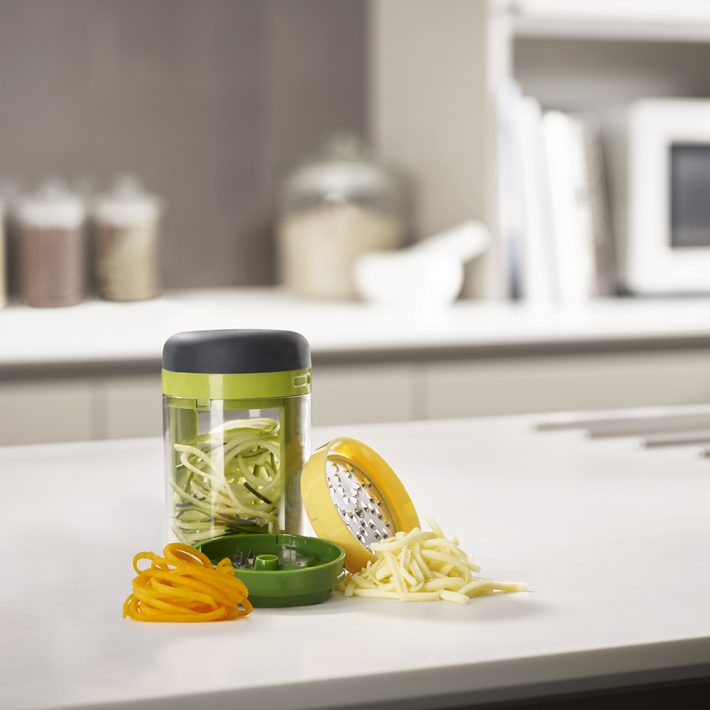 Uno spiralizzatore di verdure con verdure. Sono visibili noodles di zucchine e carote. Sono presenti anche una grattugia gialla e parti verdi.